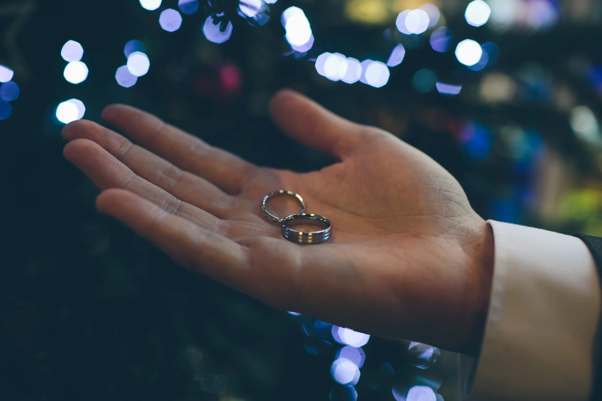 A person's hand holding two wedding rings against a background of blurred colorful lights.