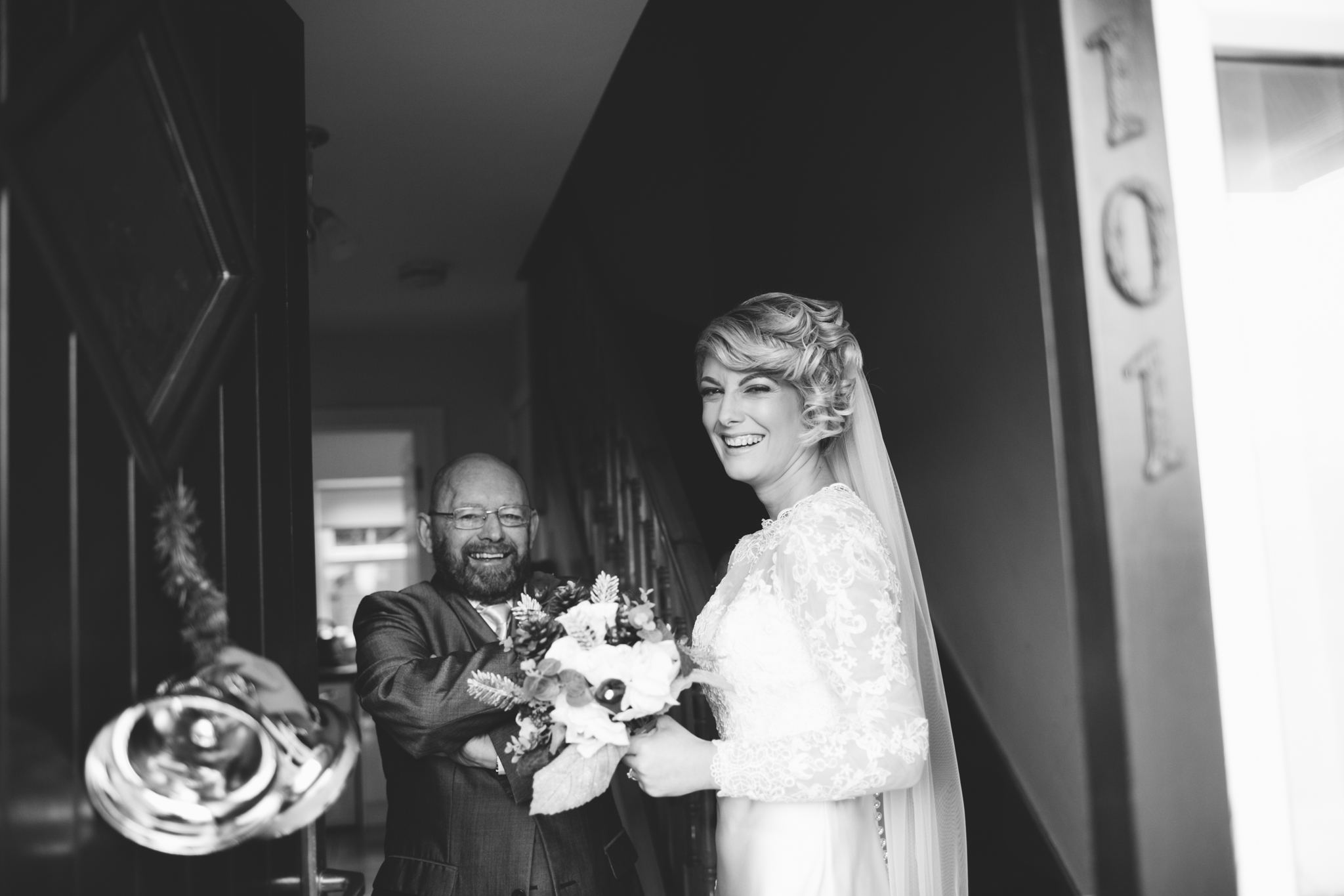 A woman in a wedding dress smiling and holding a bouquet, standing inside a home, with a man in a suit smiling and holding the bouquet toward her.