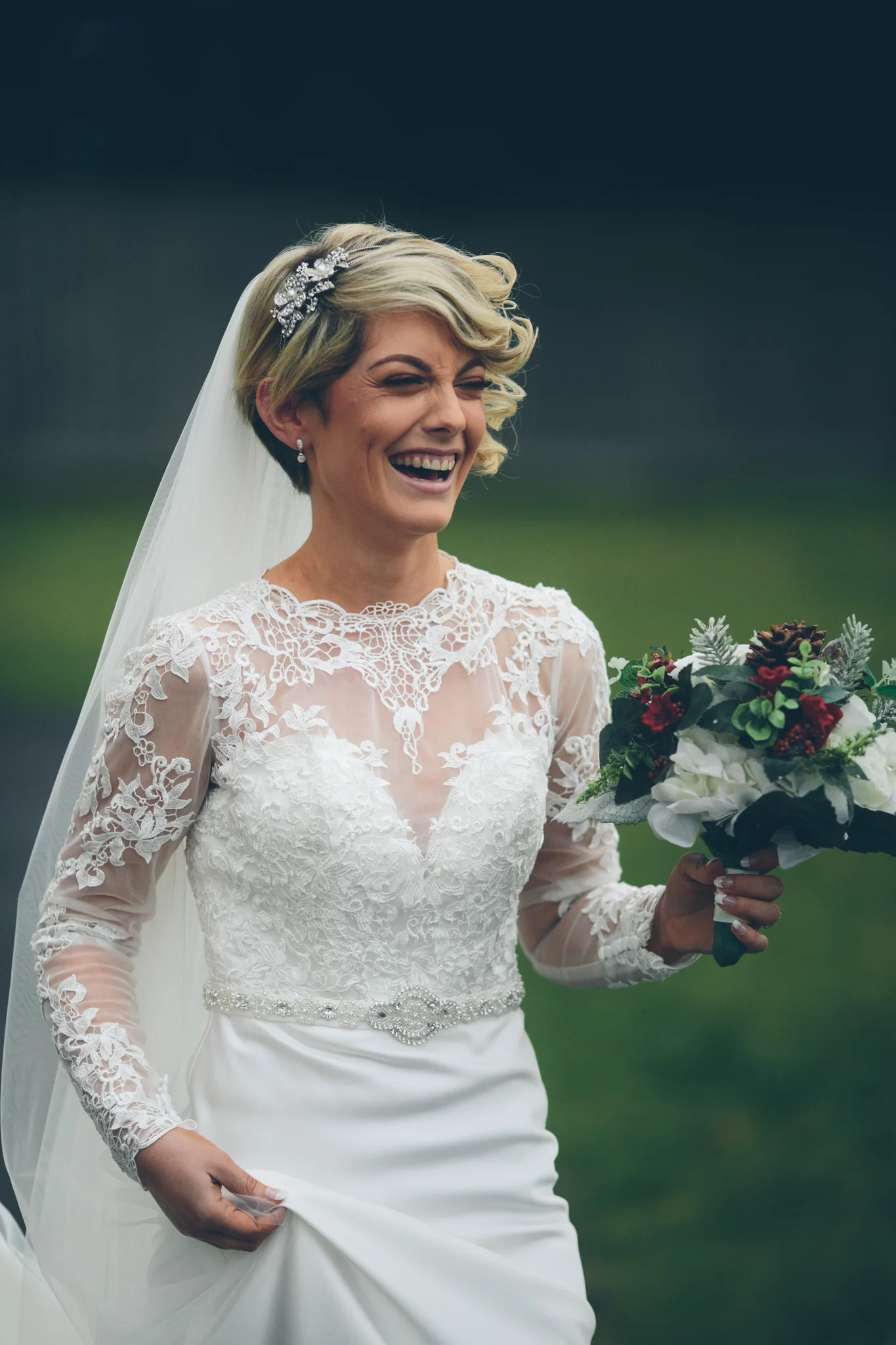 A joyful bride outdoors in a wedding dress with lace details, holding a bouquet of flowers, smiling.