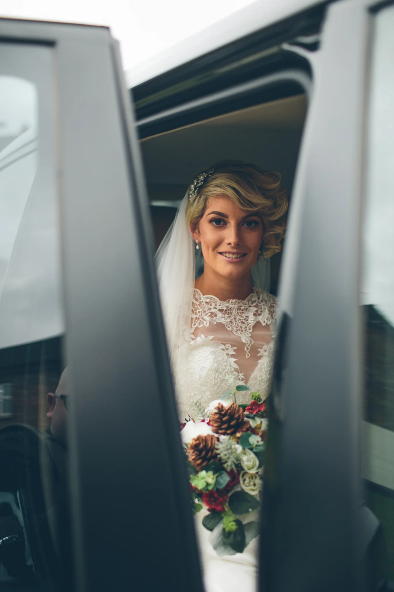 Bride looking in mirror, wearing white lace wedding dress and veil, holding bouquet, smiling.