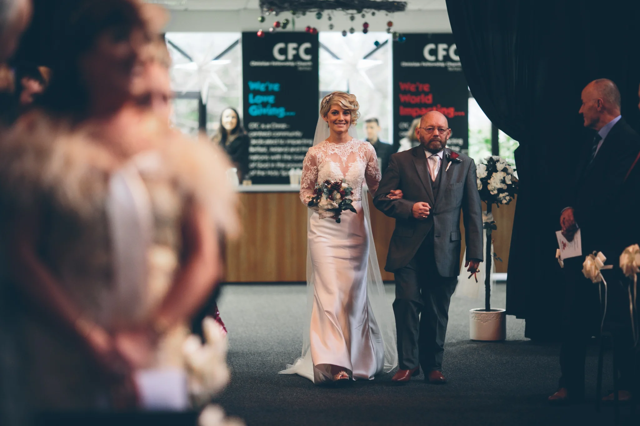 A bride walking down the aisle with her father at a wedding ceremony, surrounded by guests. The bride is wearing a white lace wedding dress and holding a bouquet.