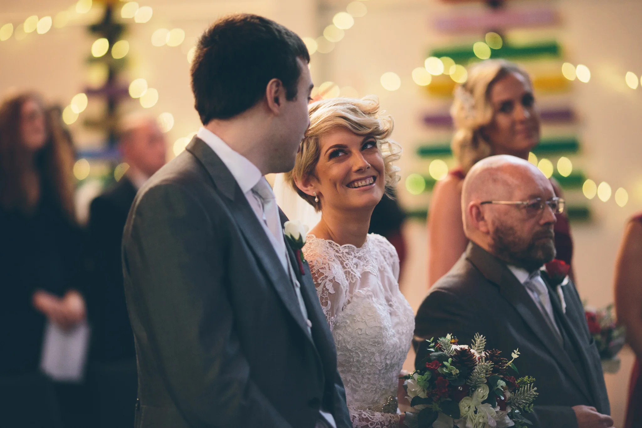 Bride and groom exchanging vows during wedding ceremony, surrounded by guests, with festive decorations and lights in the background.