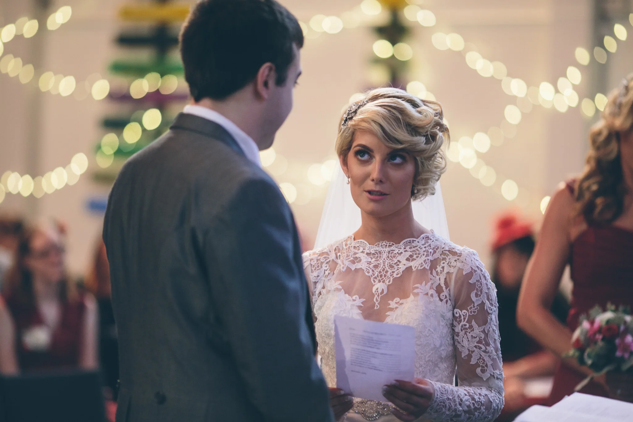 A bride and groom exchange vows or rings during their wedding ceremony in a decorated venue with string lights.