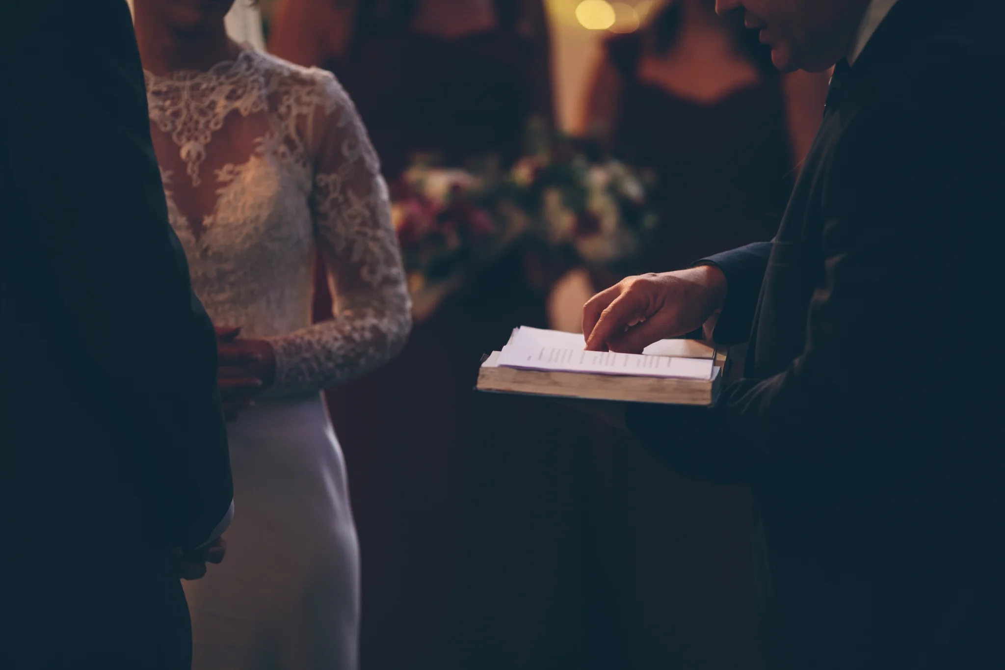 Close-up of a man in a dark suit holding a small book or hymnbook during a wedding ceremony, with a woman in a white lace wedding dress standing nearby, in a softly lit room.