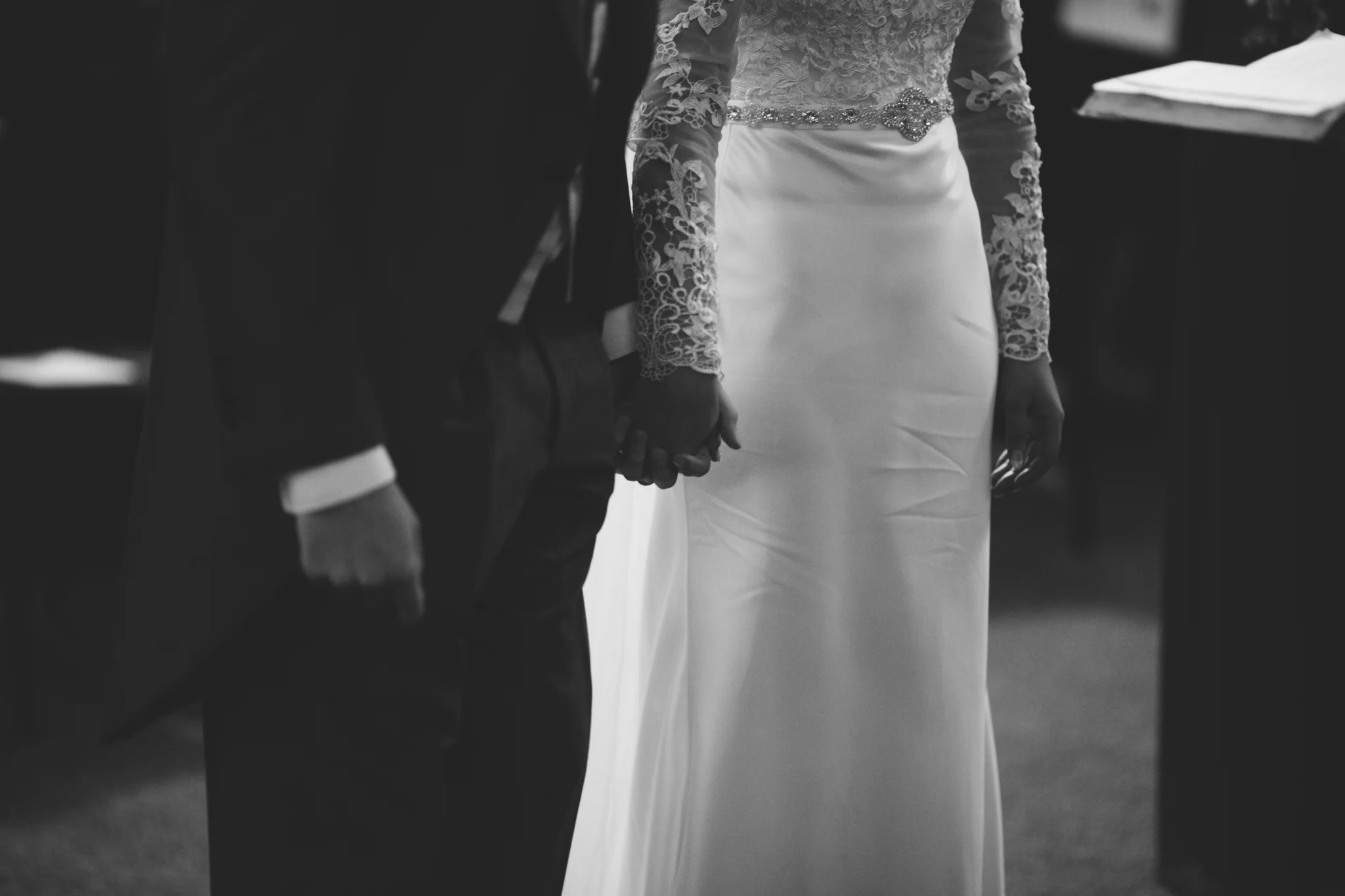 A bride and groom holding hands during their wedding ceremony, with the bride wearing a lace long-sleeve wedding dress and the groom in a suit, captured in black and white.