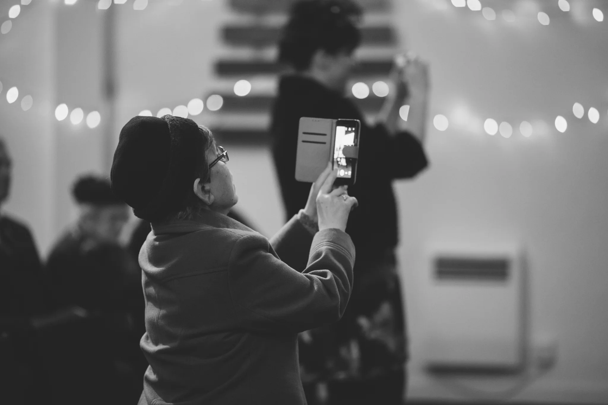 A woman wearing glasses and a beanie is taking a photo with her phone at an indoor event decorated with string lights, with people blurred in the background.