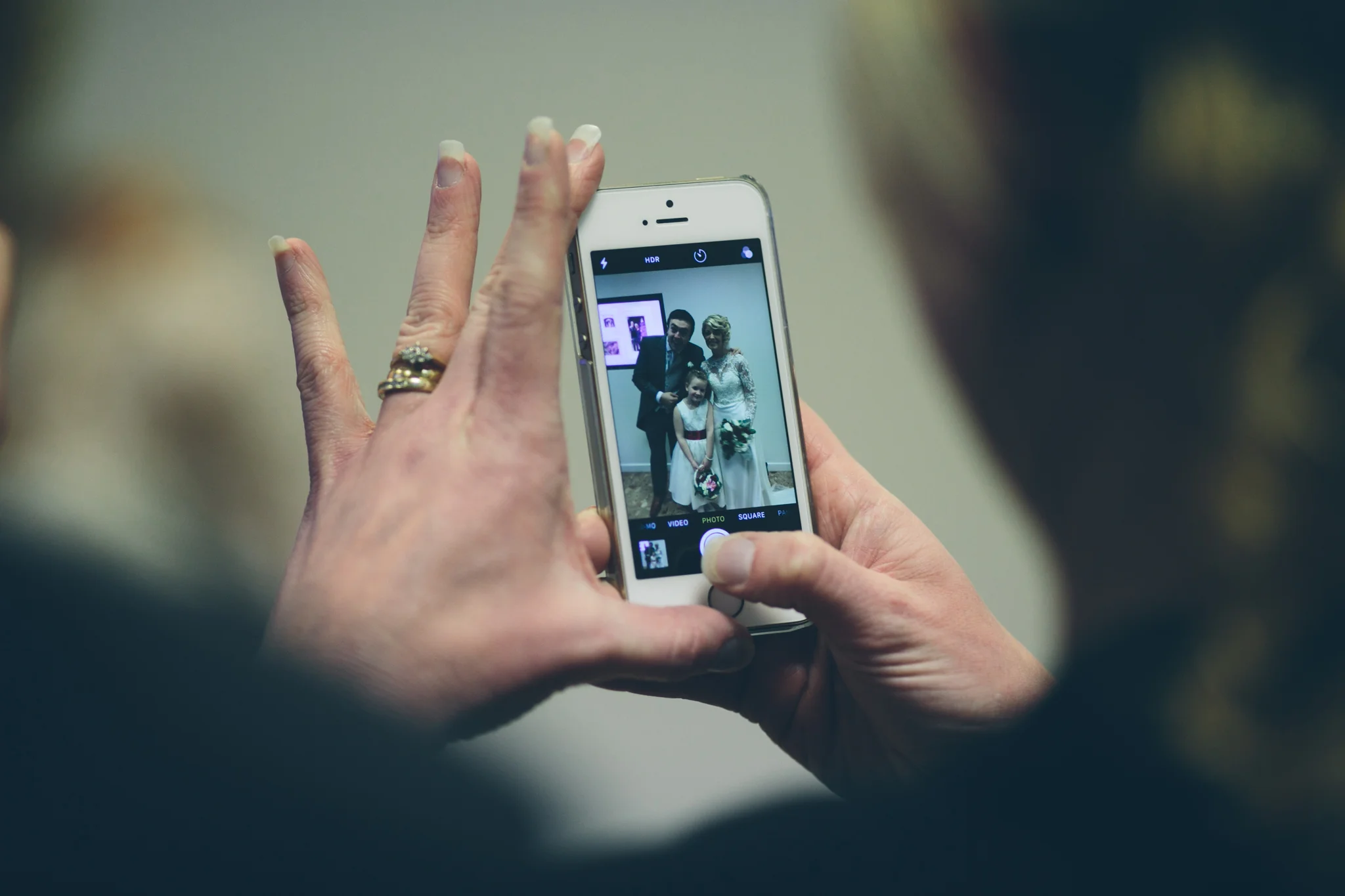 Close-up of a person taking a photo of a bride, groom, and a young girl with a smartphone in an indoor setting.