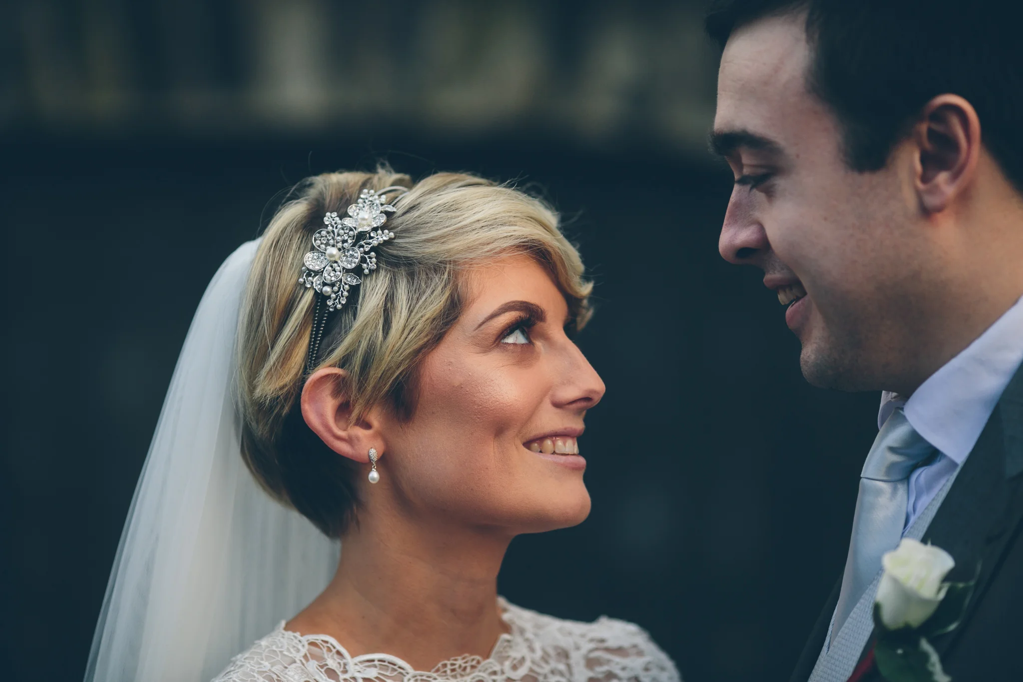 A bride and groom gazing at each other during their wedding ceremony outside, with the bride wearing a lace wedding dress and a decorative hairpiece, and the groom in a suit with a white boutonniere.