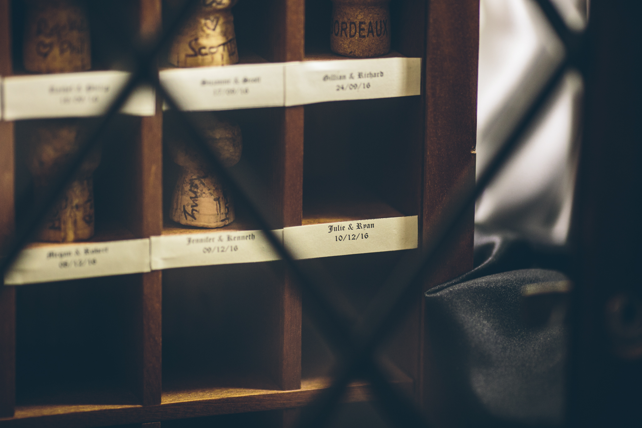 A wooden wine bottle storage box with individual compartments labeled with names and dates, viewed through a wire mesh fence.