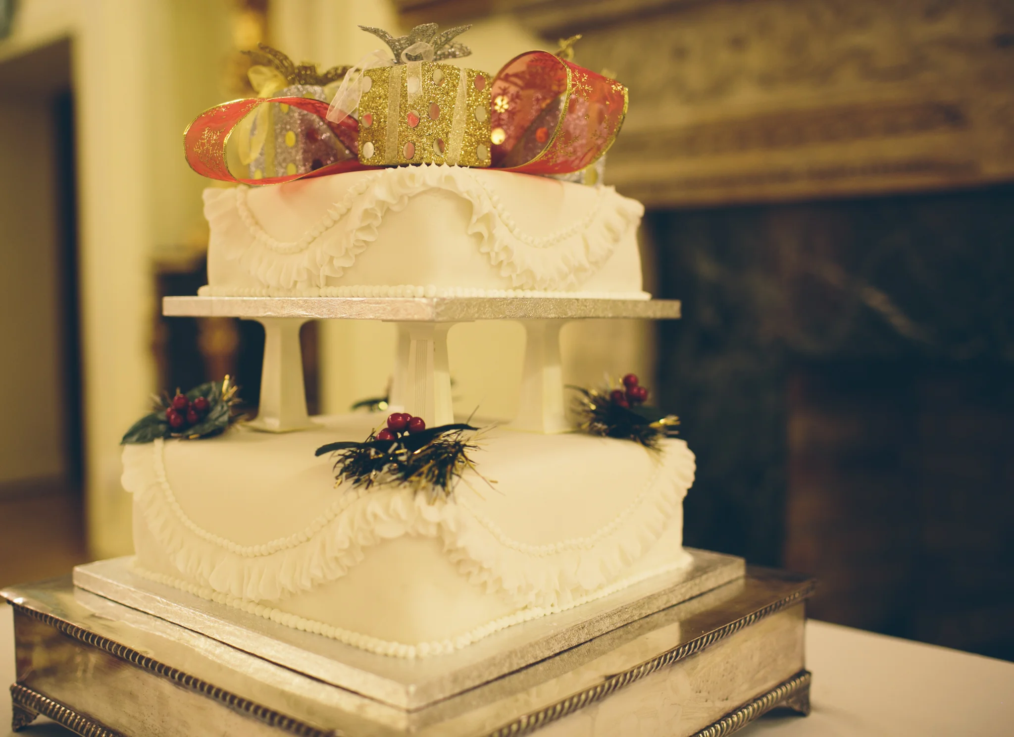 A two-tiered white wedding cake decorated with holly leaves and red berries, topped with a large decorative bow and gold gift box, on a silver cake stand.