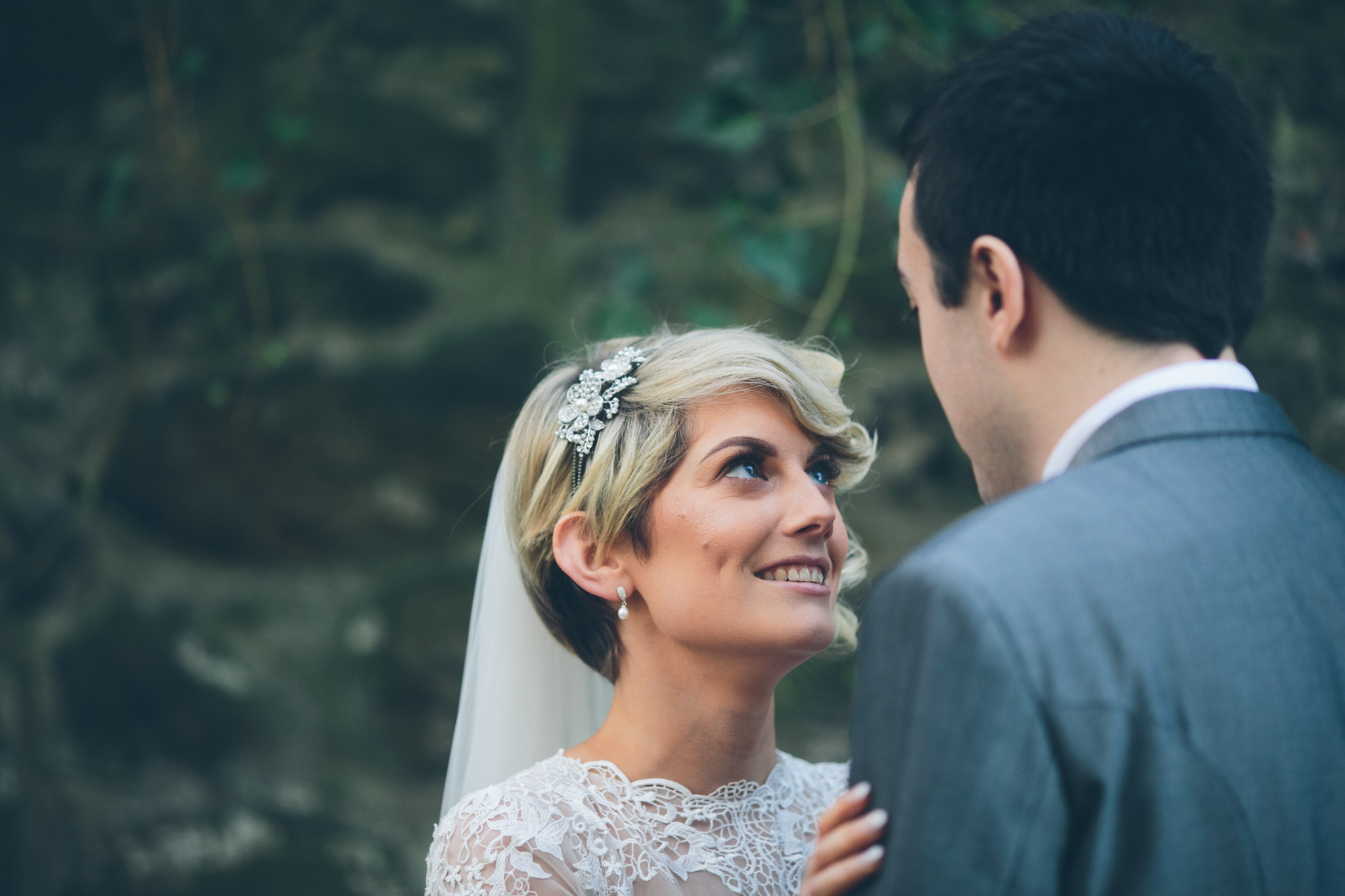 A bride with short blonde hair and wearing a lace wedding dress looks into the eyes of a groom with dark hair in a gray suit near a body of water with rocks and green foliage in the background.