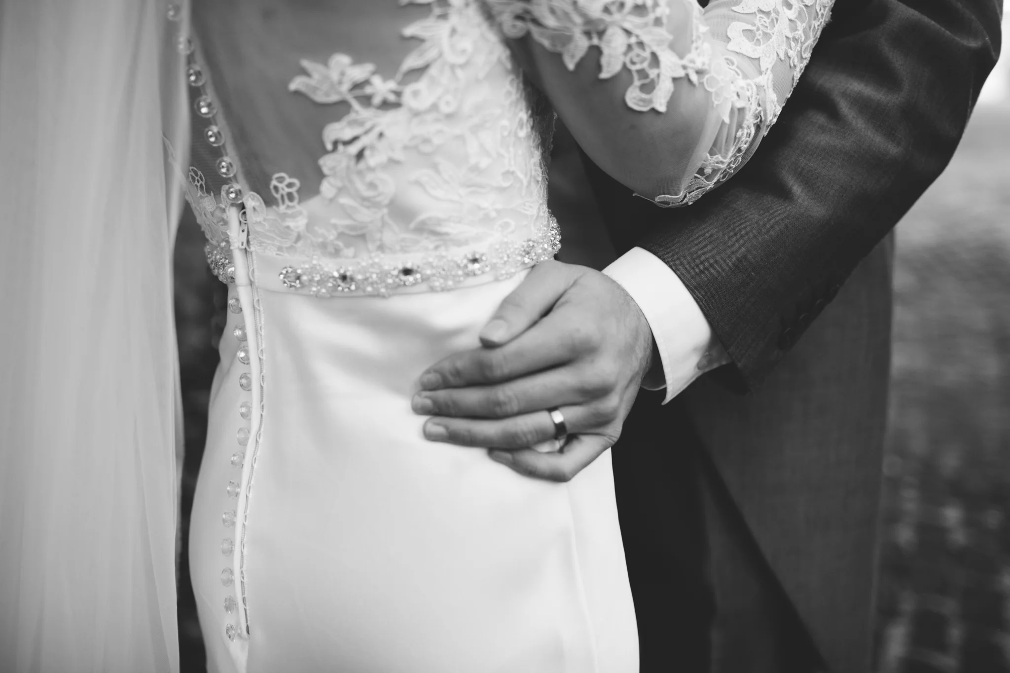 Close-up of a bride and groom holding hands during their wedding, focusing on the bride's wedding dress with lace details and buttons, and the groom's hand with a wedding ring.