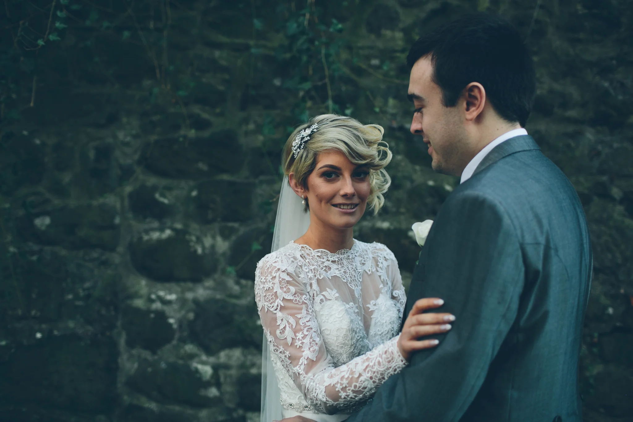 A bride and groom face each other outdoors, holding hands, with a stone wall in the background.