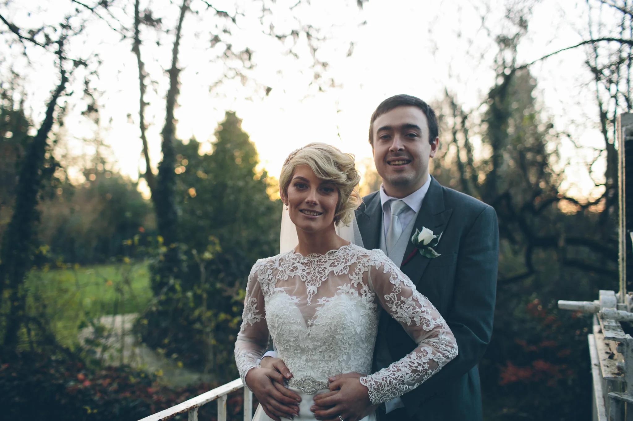 A bride and groom in wedding attire standing on a bridge outdoors, with trees and a sunset in the background.