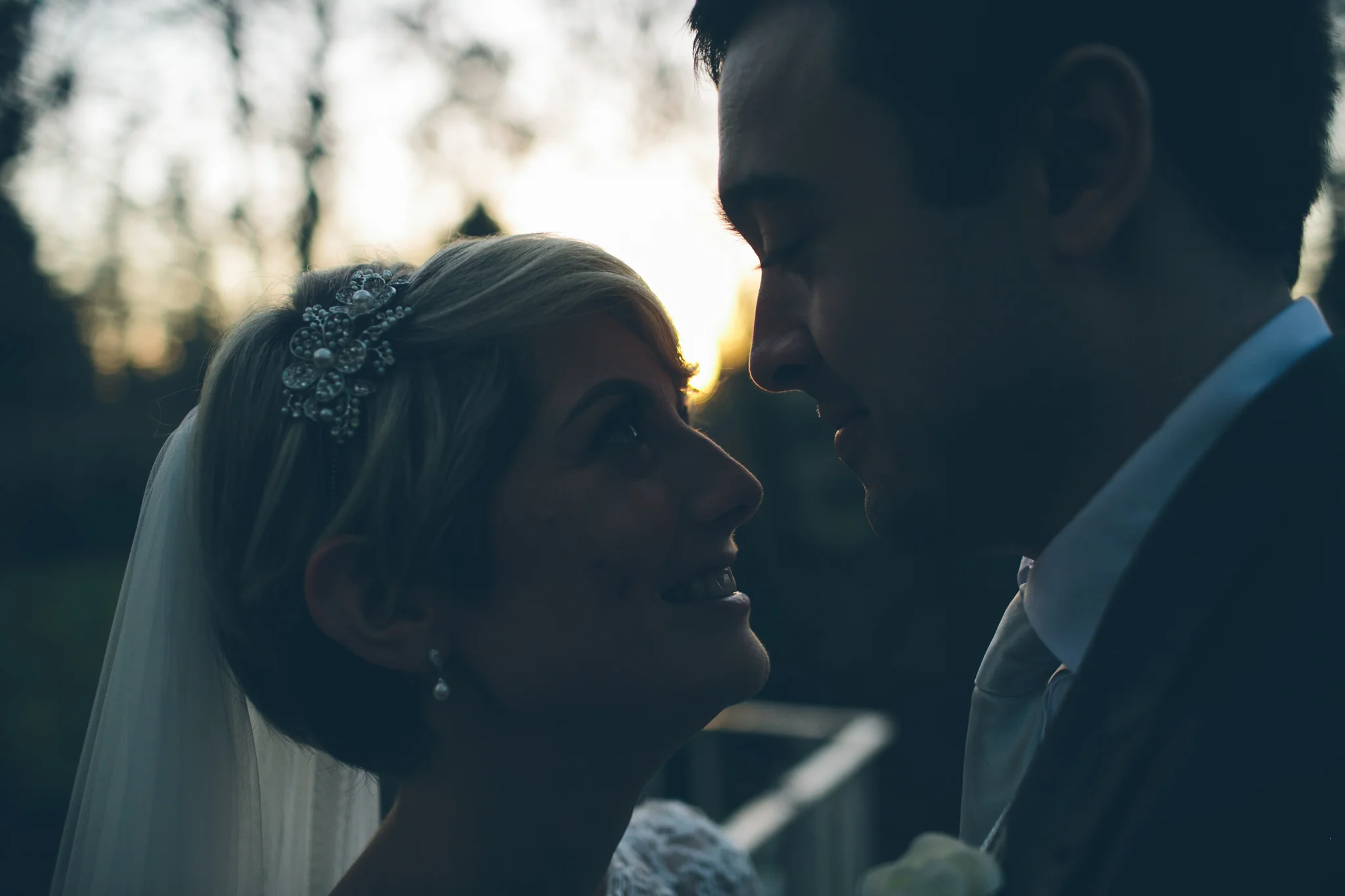 A bride and groom face each other outdoors during sunset, with their foreheads touching and smiling at each other.