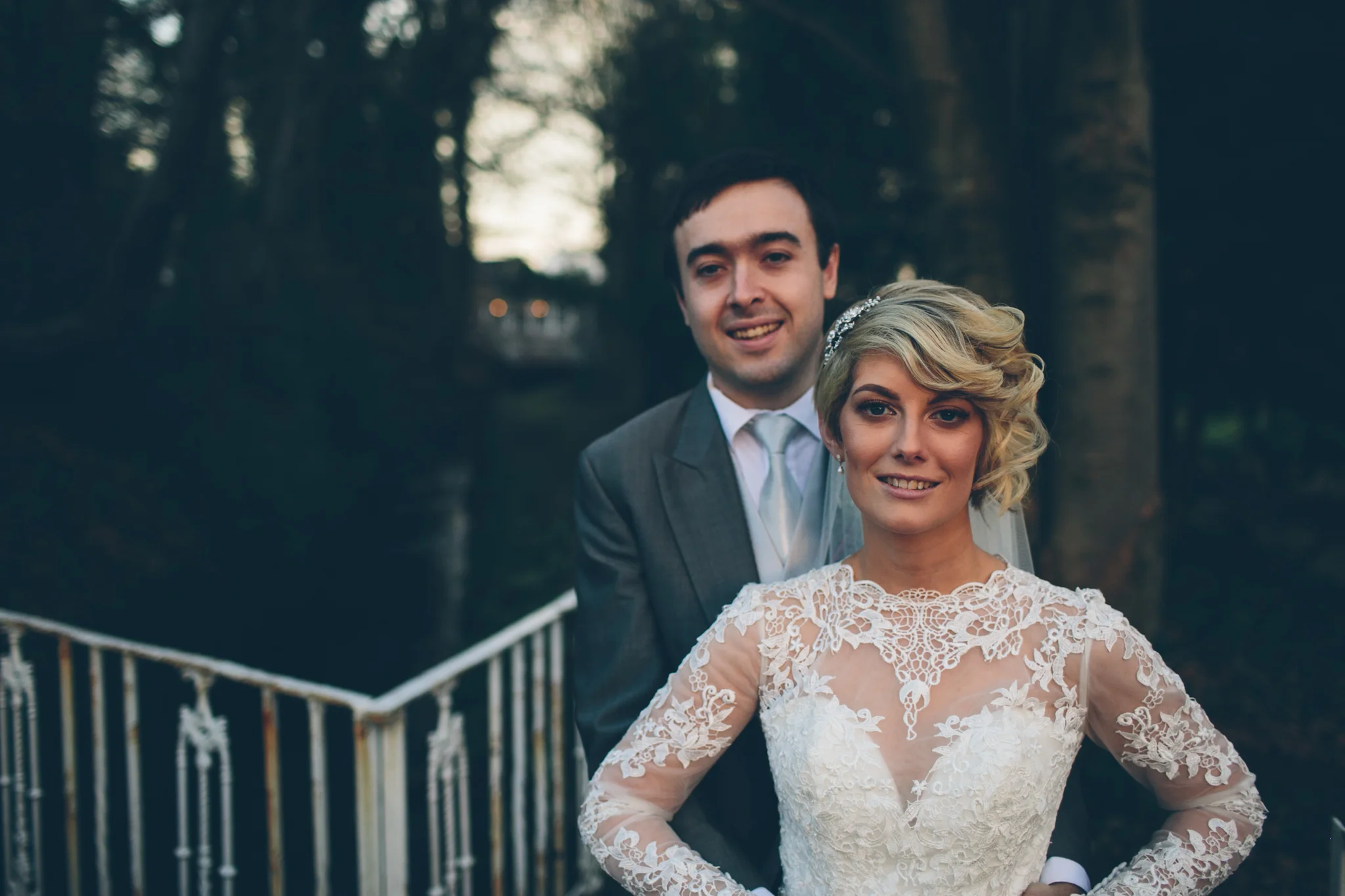 A bride and groom standing outdoors near a white railing, smiling at the camera. The bride is wearing a lace wedding dress with long sleeves and has short, wavy blonde hair. The groom is wearing a gray suit with a white shirt and tie, with short dark