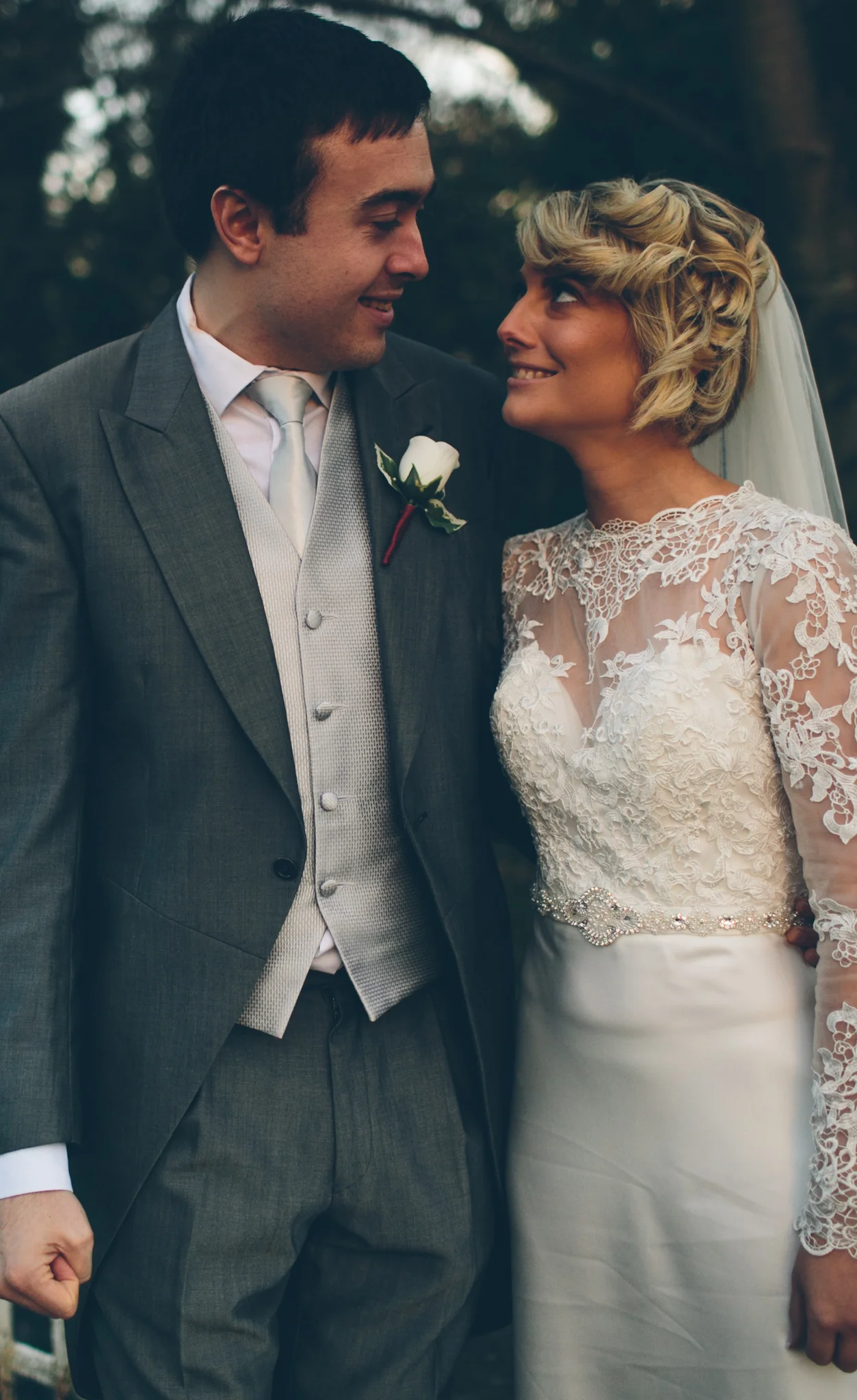 A bride and groom look at each other and smile during their wedding. The groom is wearing a gray suit with a white shirt, tie, and vest, and has a white rose boutonniere. The bride is wearing a white lace wedding dress with long sleeves, a decorative