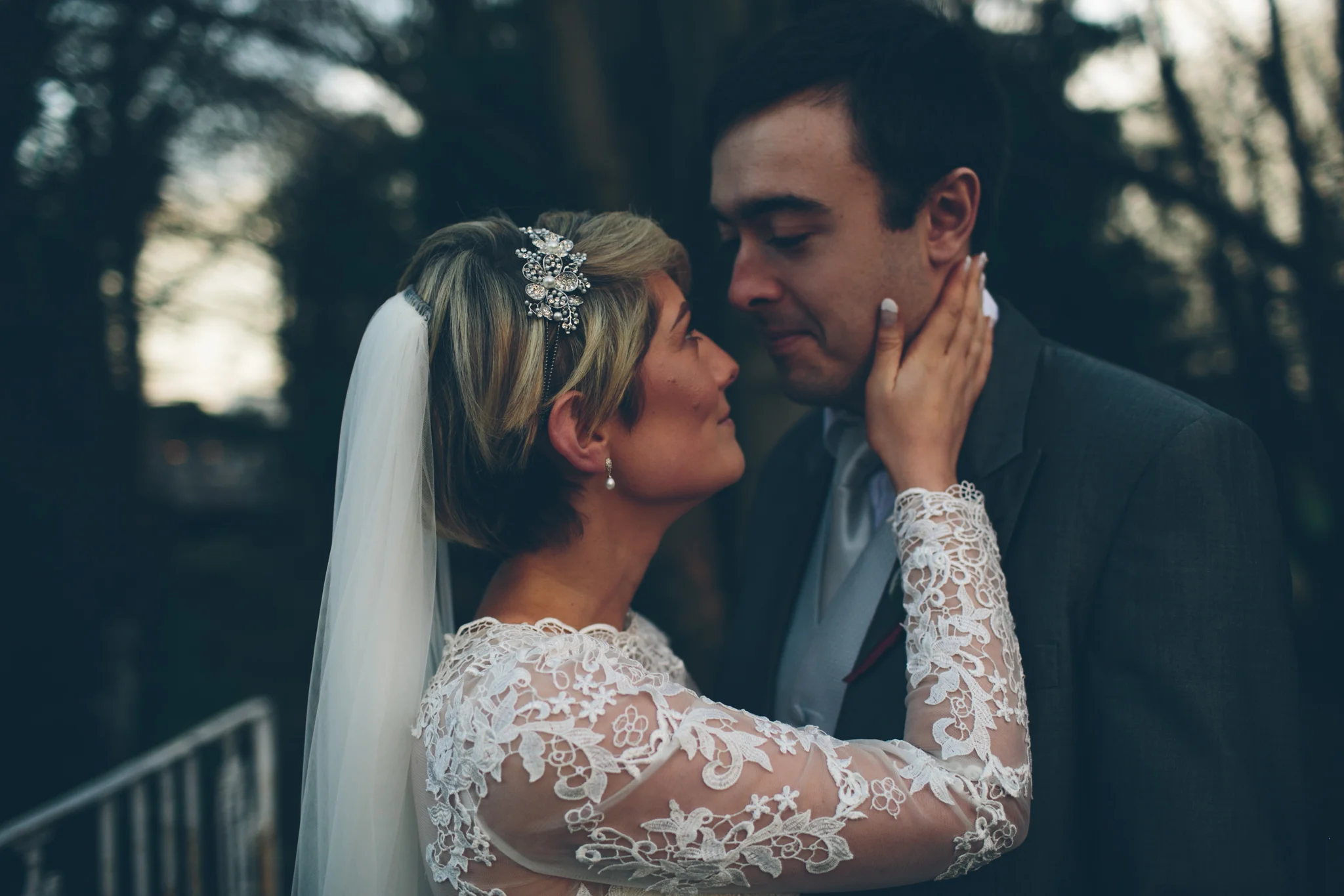 A bride and groom share an intimate moment outdoors, standing very close with foreheads touching, amidst blurred trees and natural scenery.