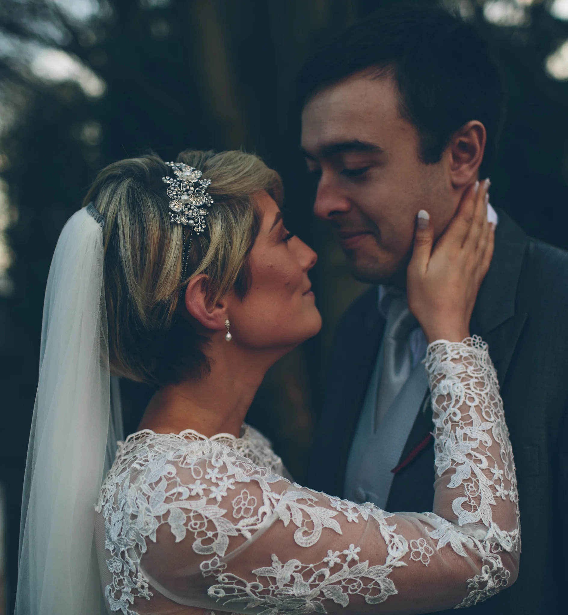 A bride and groom with their foreheads touching, the bride holding the groom's face, outdoors during the evening.