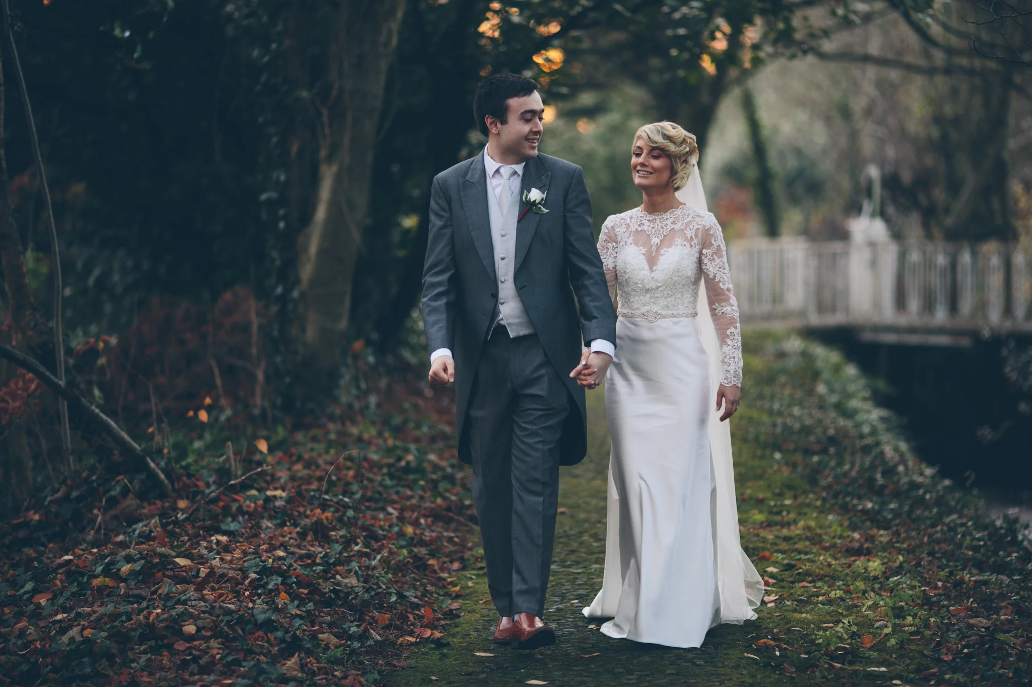 A bride and groom walking hand in hand outdoors on a wooded path during their wedding day. Dunadry Hotel