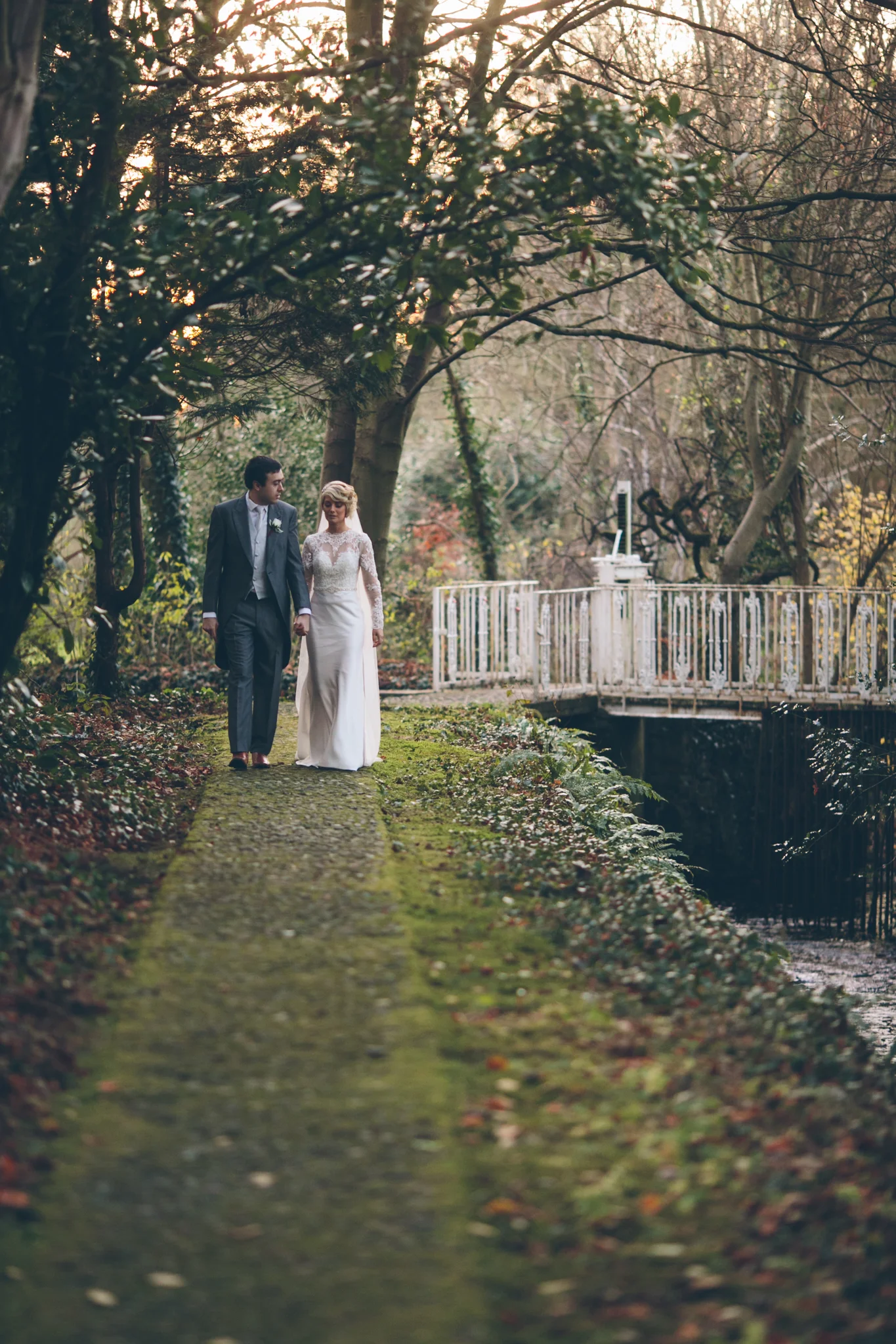 A bride and groom walking hand in hand along a mossy, narrow garden path surrounded by trees.