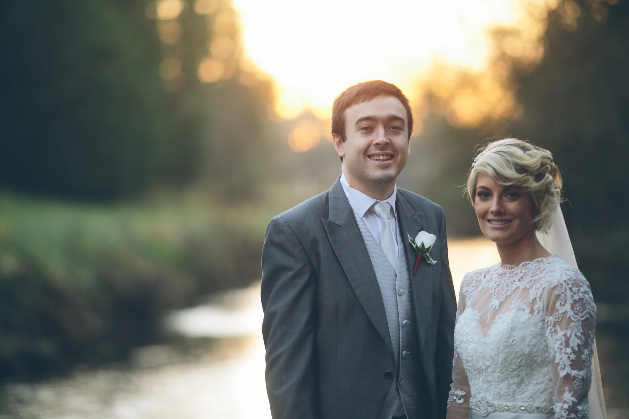A newlywed couple stands outdoors near a body of water during sunset, smiling at the camera. The groom wears a gray suit, and the bride wears a lace wedding dress with a veil. Dunadry Hotel