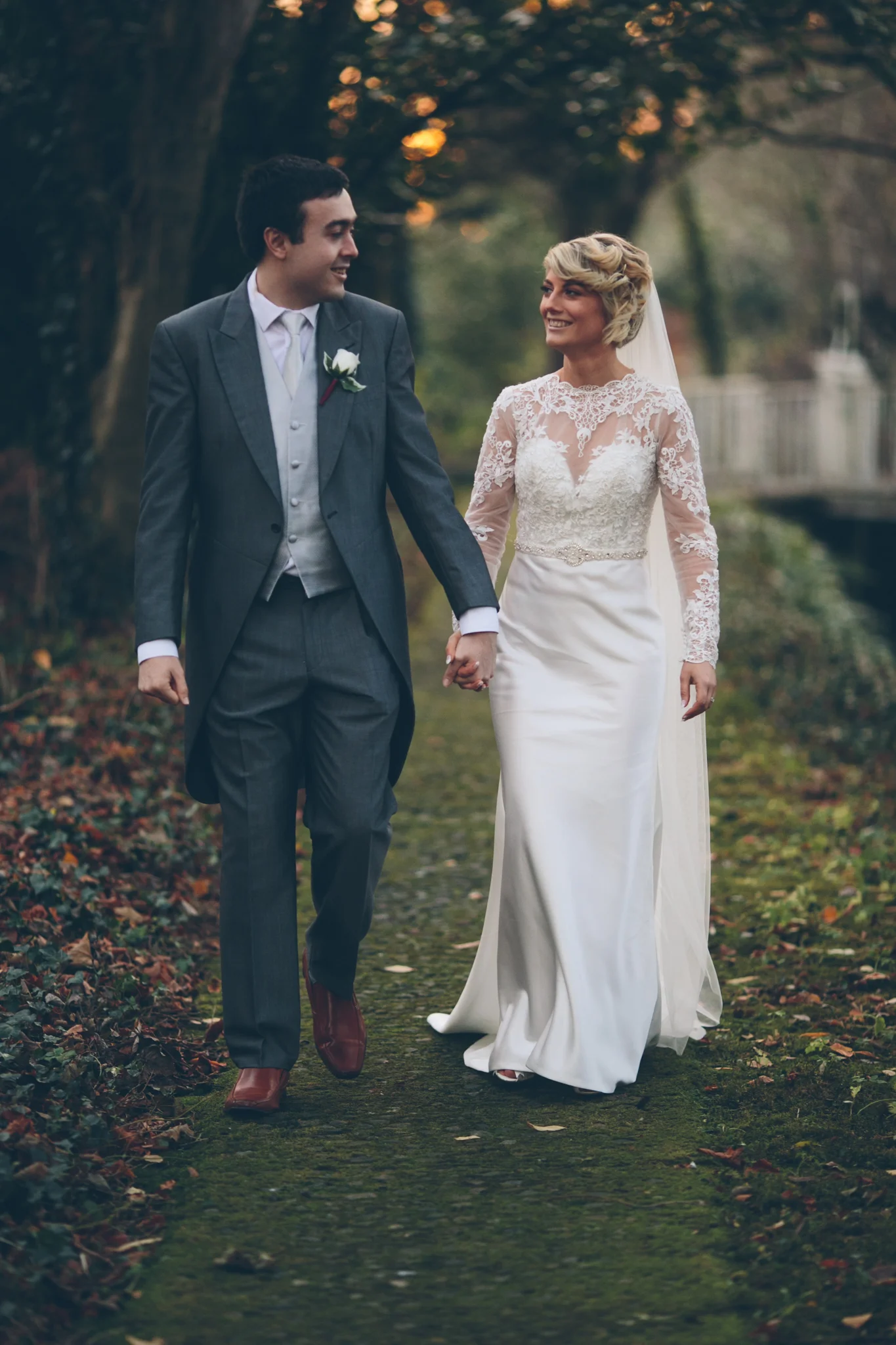 A bride and groom walking hand in hand outdoors during sunset, surrounded by trees and fallen leaves.