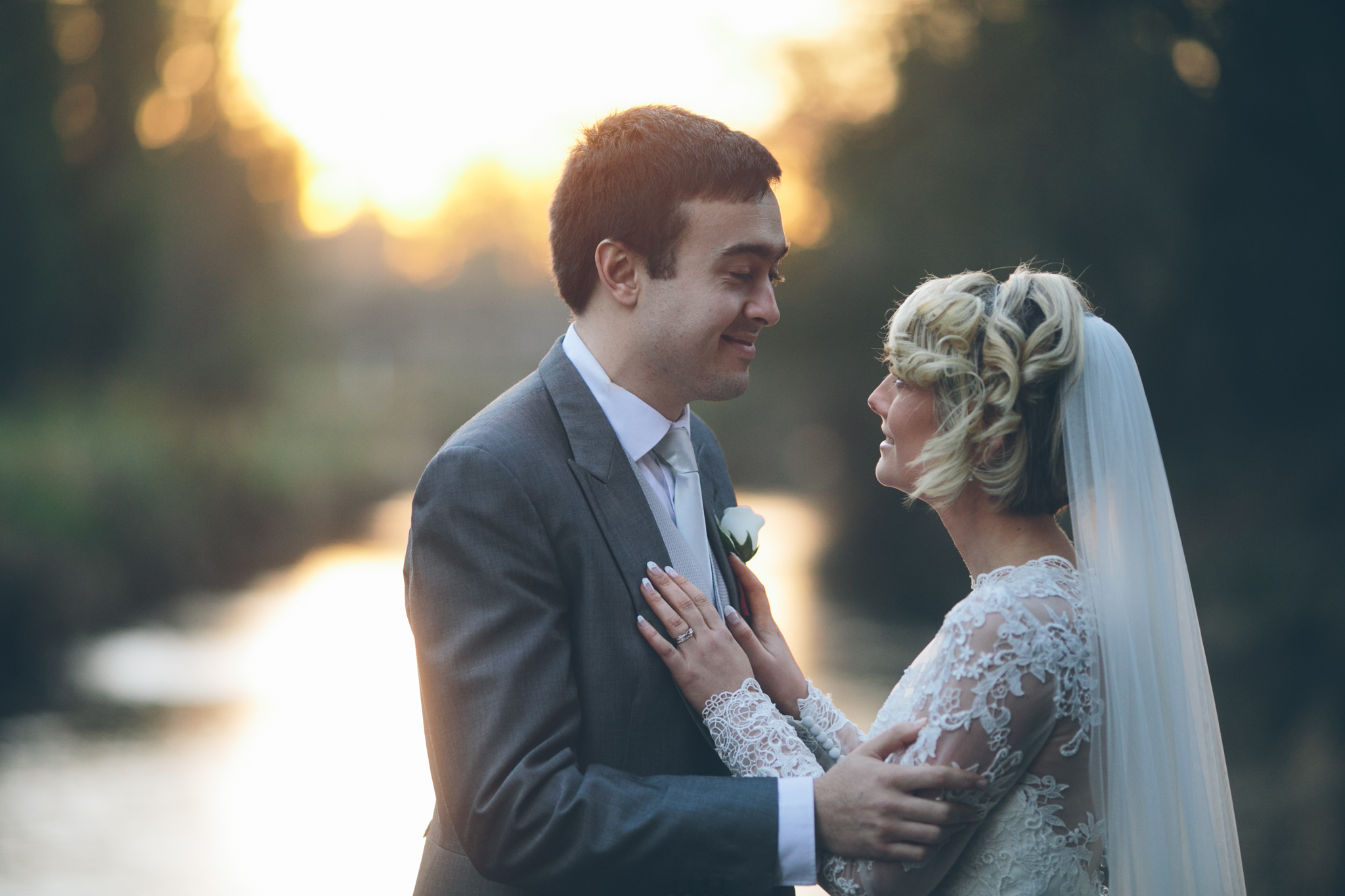 A bride and groom standing close together outdoors at sunset, facing each other tenderly, with the bride’s hands on the groom’s chest, near a body of water and surrounded by nature.