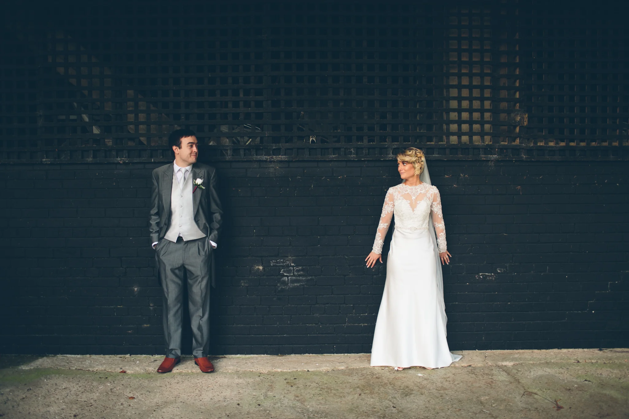 A bride and groom stand apart against a black brick wall, looking at each other. The groom wears a gray suit with a white shirt, gray tie, and a white pocket square. The bride wears a white lace wedding dress with long sleeves and a veil.