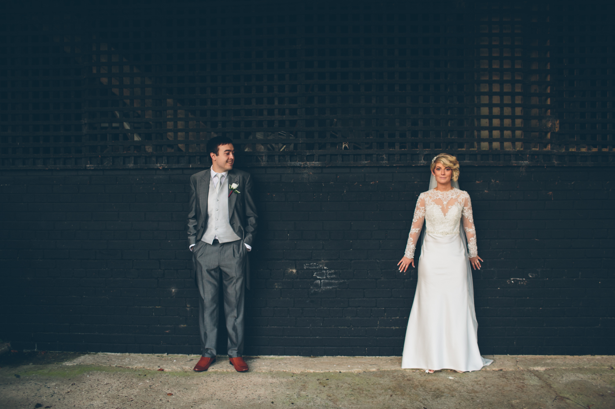 Bride in a white wedding dress and groom in a gray suit stand against a black brick wall with a trellis above, facing each other. Dunadry Hotel.