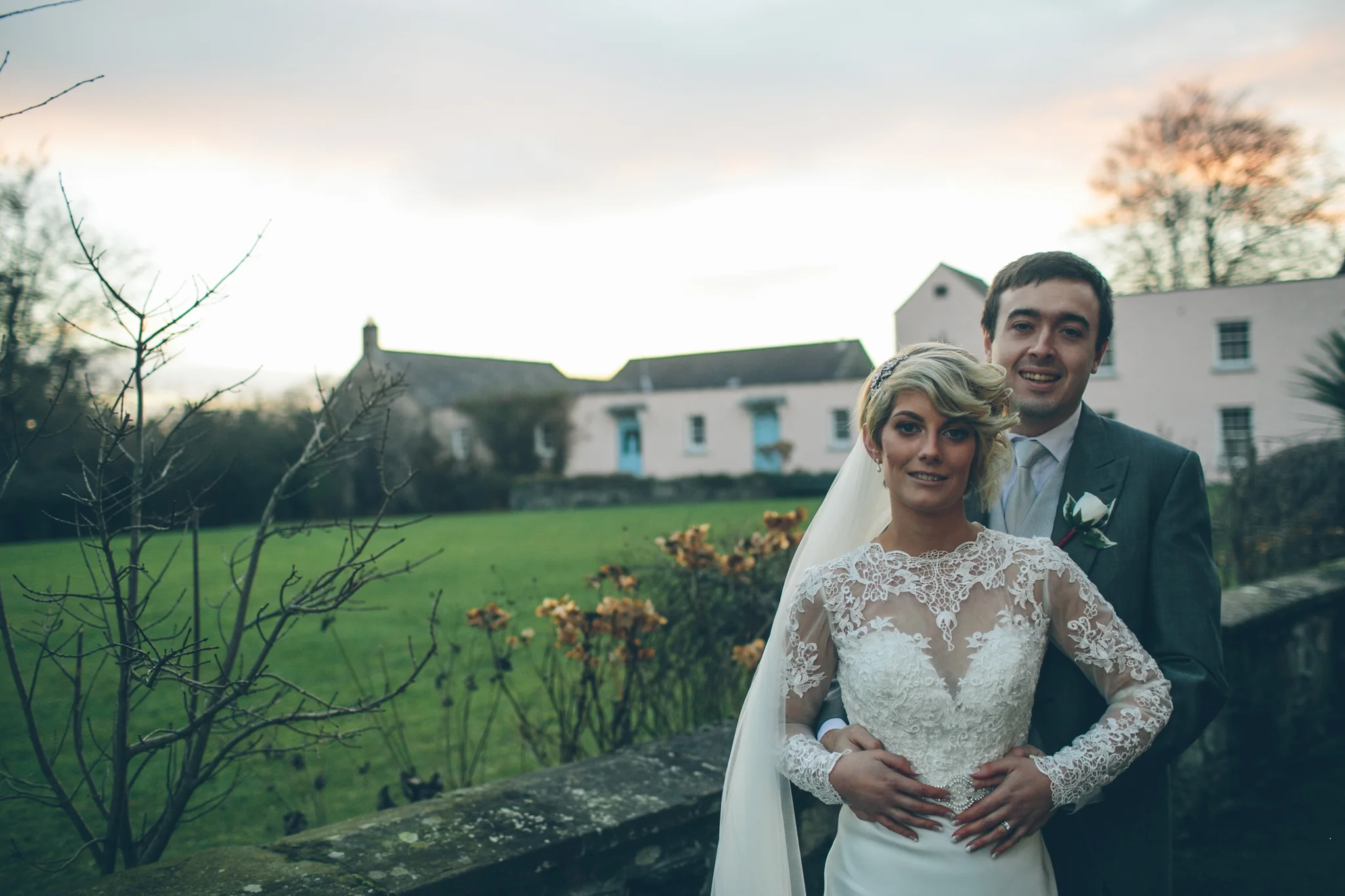 Bride and groom standing outdoors in front of a stone wall at sunset, with trees and white houses in the background, during wedding photoshoot. Dunadry Hotel.