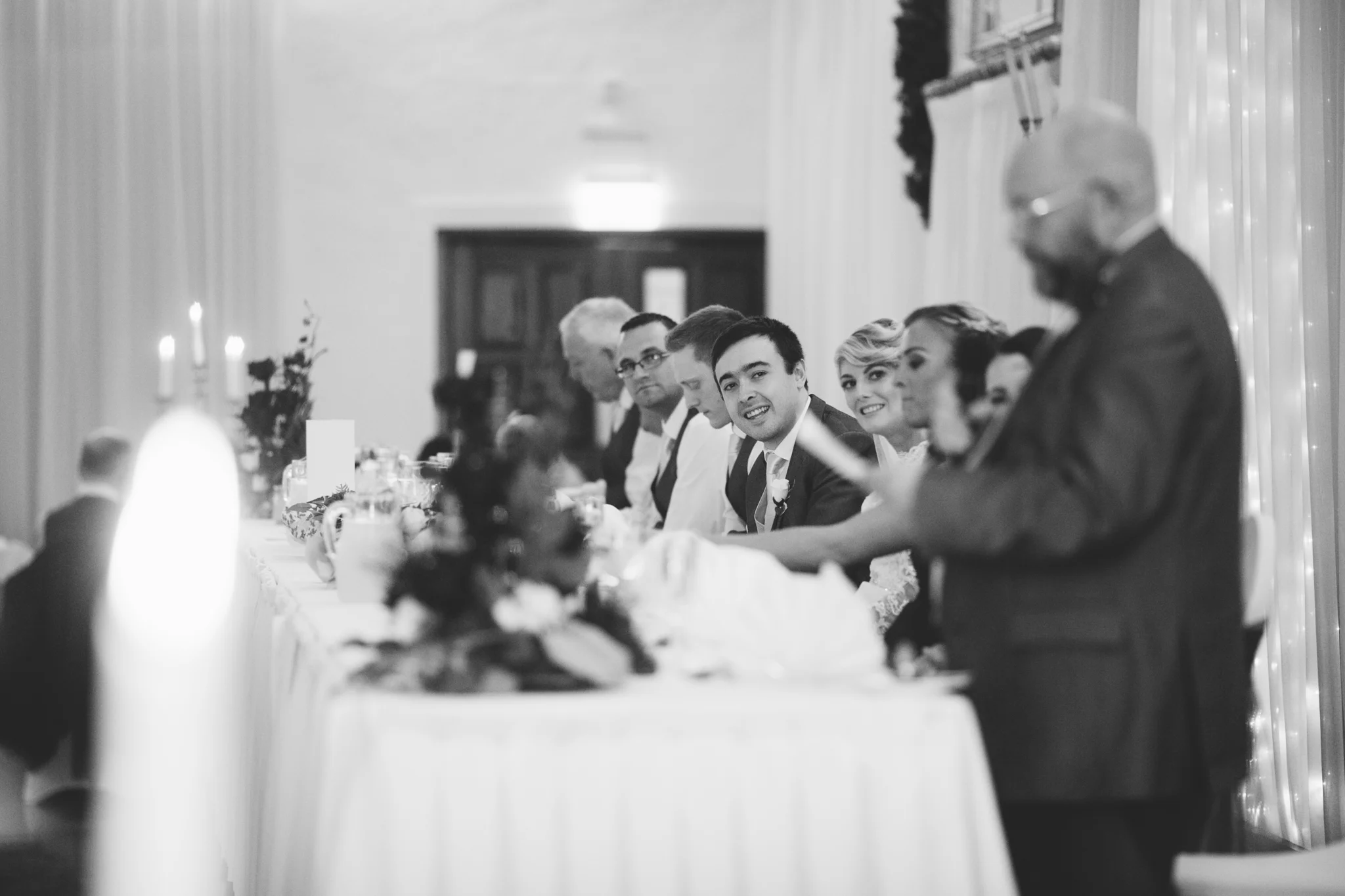 People seated at a wedding reception table, with one person speaking and others listening, in an elegant decorated room.