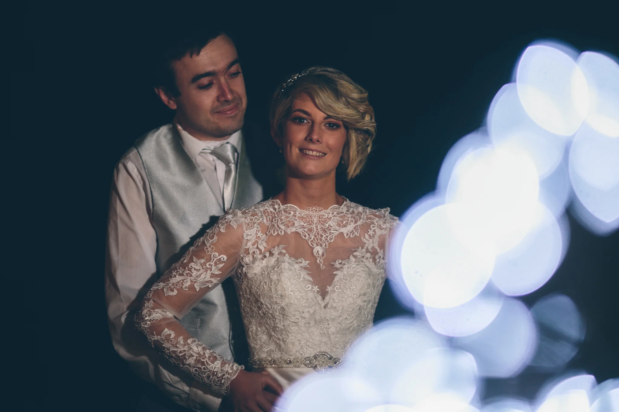 A bride in a lace wedding dress smiling, standing in front of a groom in a suit, with blurred lights in the foreground.