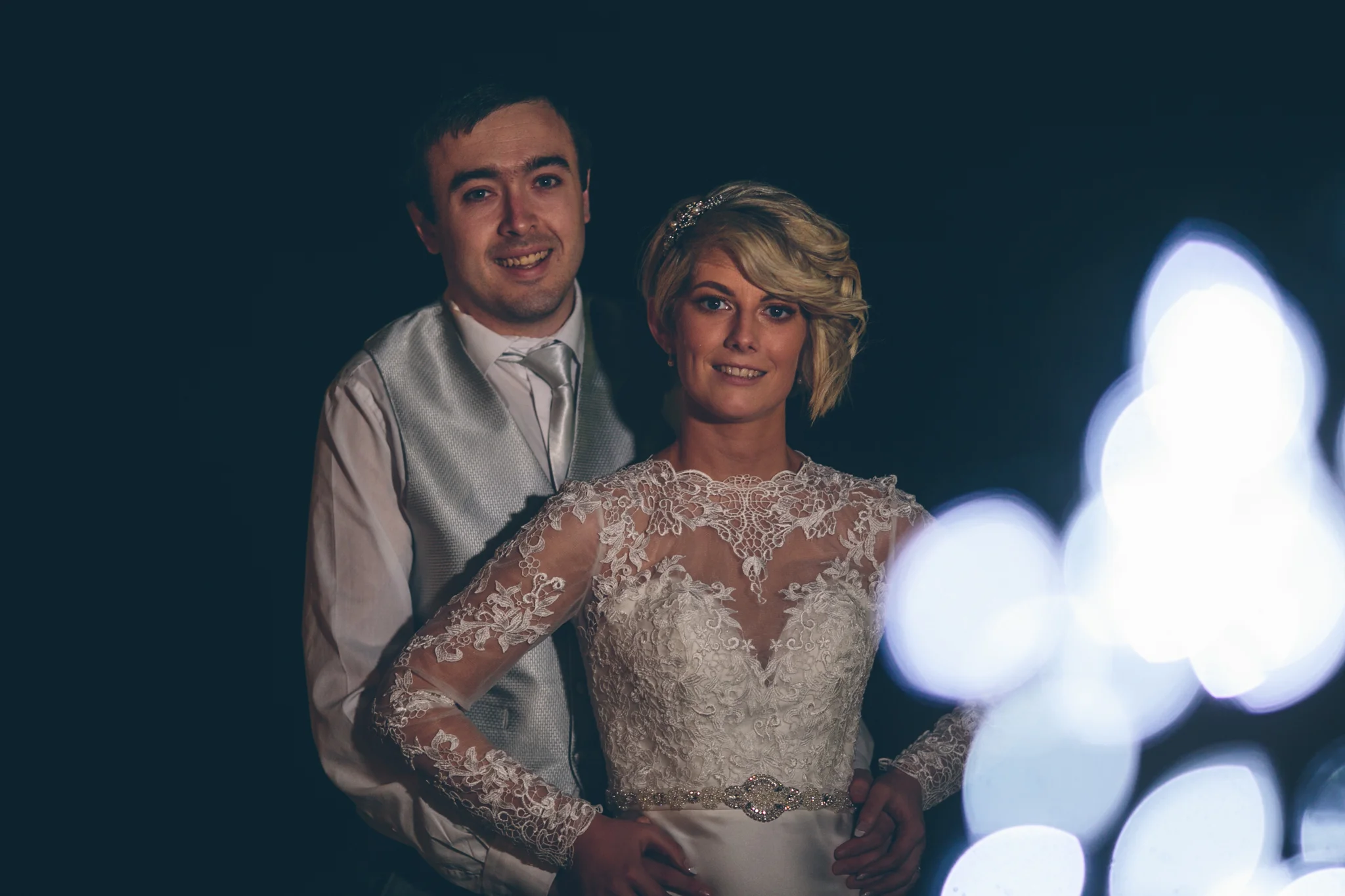 A bride and groom posing together at a wedding, with the bride in a lace wedding gown and the groom in a suit with a vest and tie, against a dark background with bright bokeh lights in the foreground.