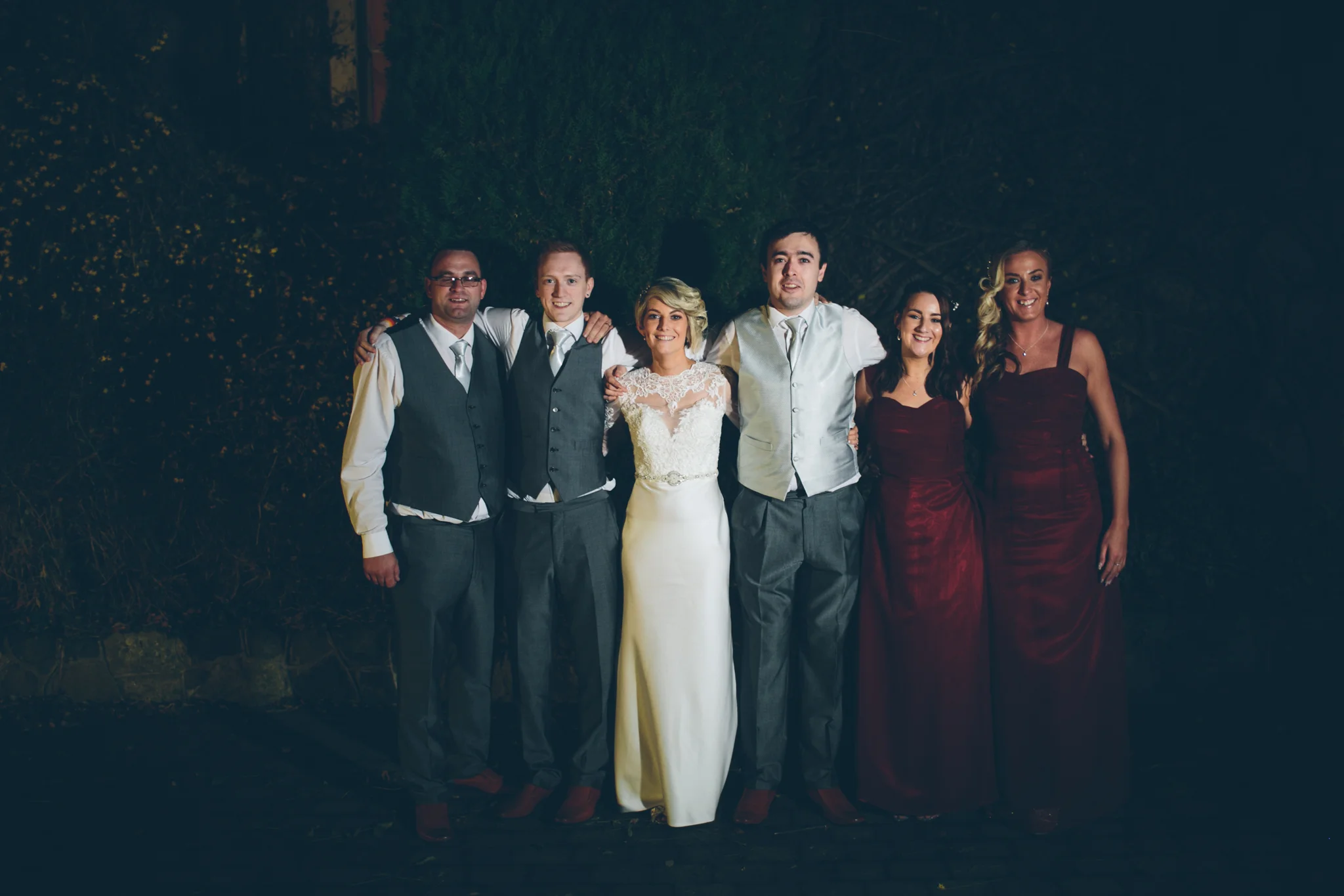 Group of wedding guests standing together outdoors at night, smiling, dressed in formal attire, with a bride in a white gown in the center. Bridal Party. Dunadry Hotel.