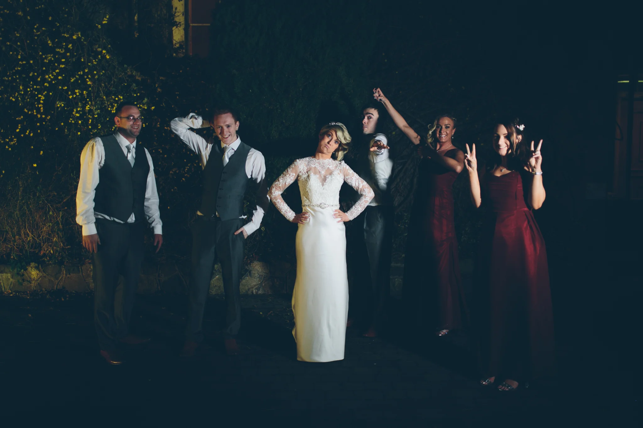 Group of seven people dressed in formal attire at night, standing outdoors against bushes and trees. The woman in the center wears a white lace dress, and the others wear suits and dresses, some making peace signs and smiling.