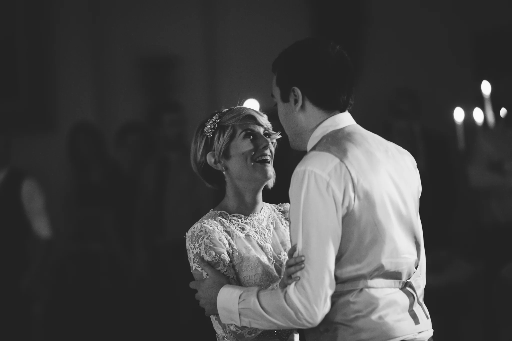 A black and white photo of a couple dancing and smiling at a formal event, with blurred background featuring other people and candlelight.