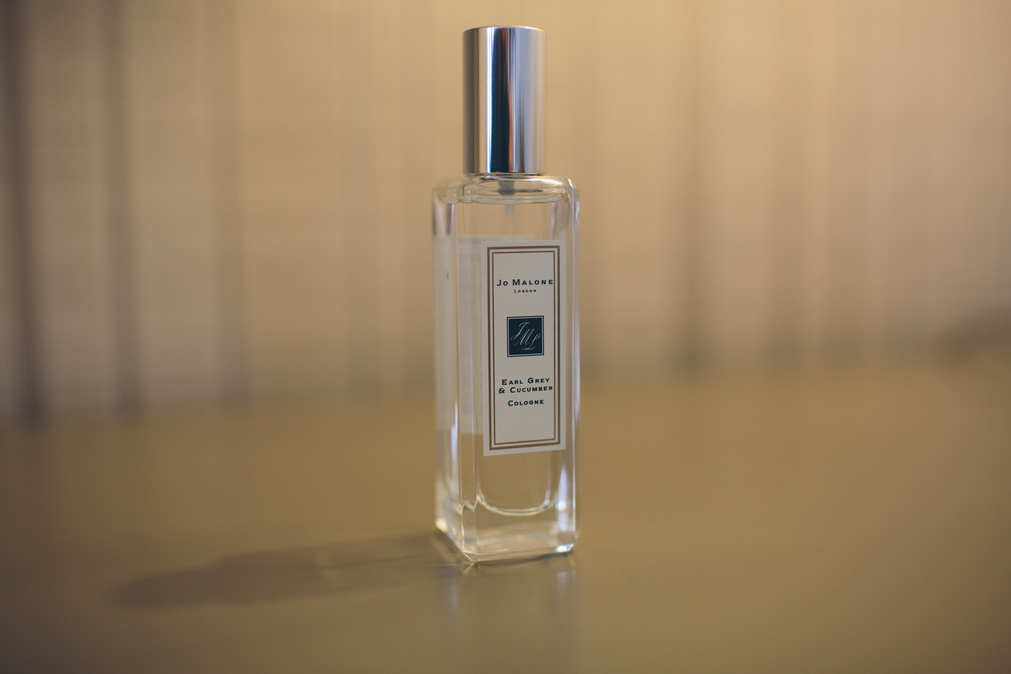 A clear rectangular glass bottle with a silver cap, labeled Jo Malone London Earl Grey & Cucumber cologne, standing on a beige surface with a blurred beige background.