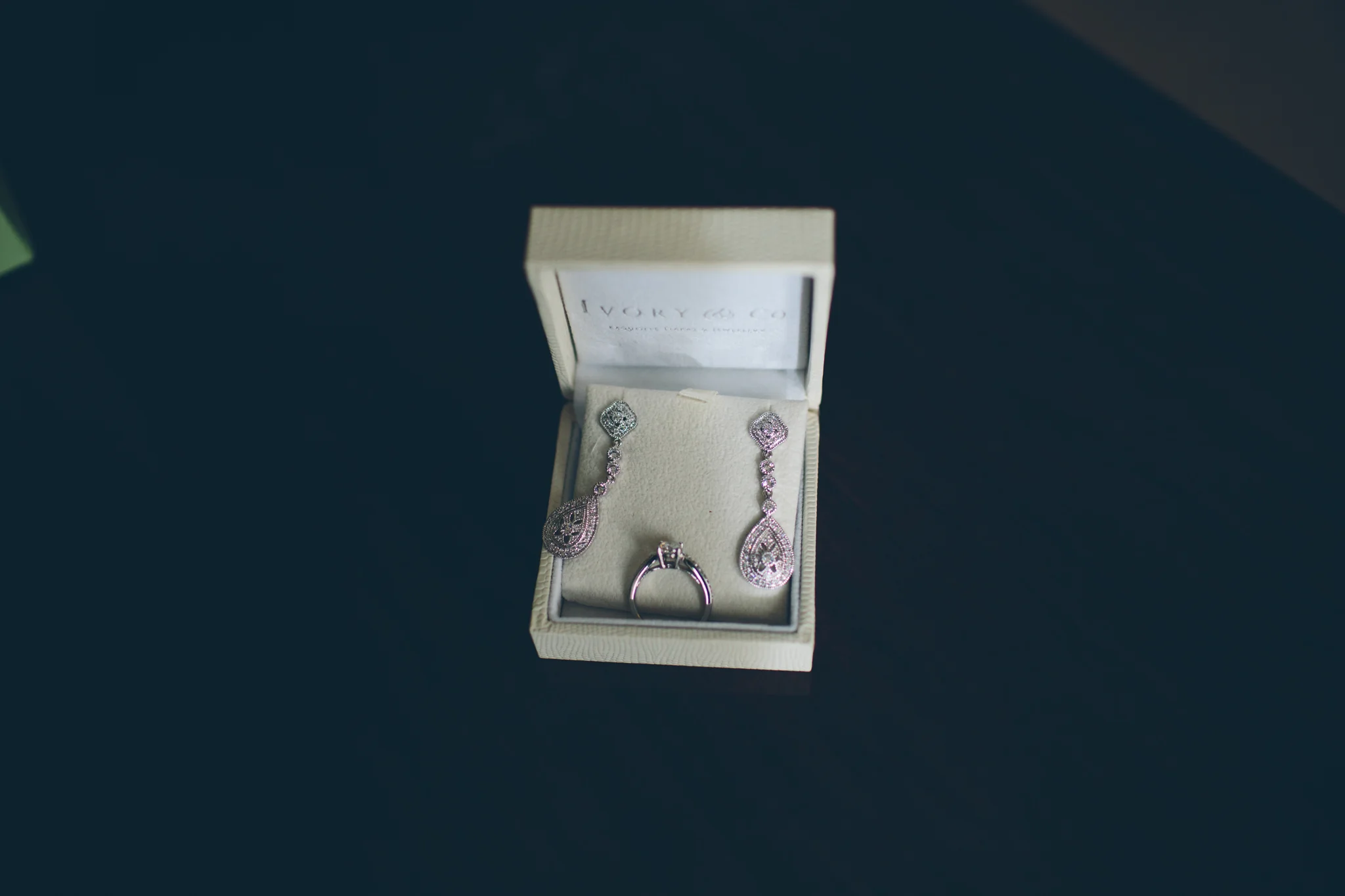 A jewelry box containing a pair of pink and clear gemstone earrings and a silver engagement ring on a dark surface.