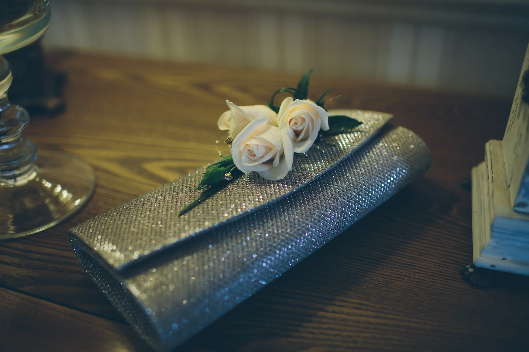 A silver, glittery clutch purse with three white roses on top, placed on a wooden surface.