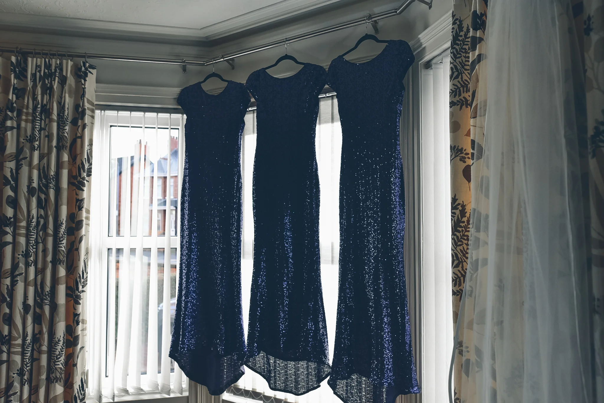 Three blue sequined evening gowns hanging on hangers in front of a window with curtains.