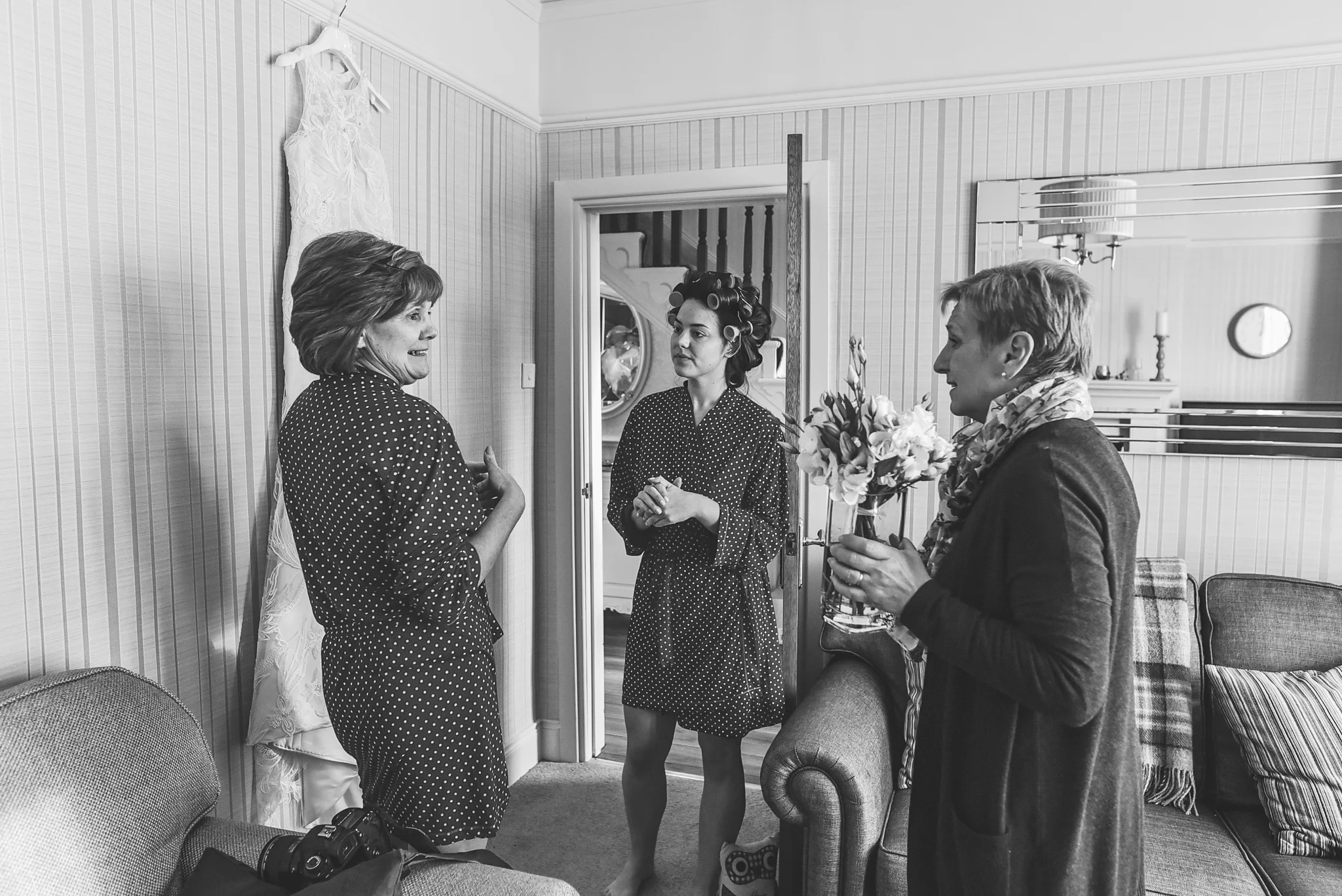 Three women in a living room, with one woman holding a bouquet of flowers, all engaged in conversation. One woman is wearing a polka dot dress and hair rollers, another in a polka dot robe, and the third in a dark coat with a scarf. There is a camera