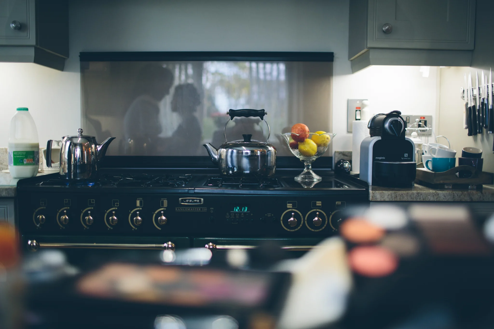 Kitchen stove with a kettle and a tea pot on top, bowl of apples and lemons, coffee machine, cups, and kitchen utensils. Reflection of two people in a window behind the stove.