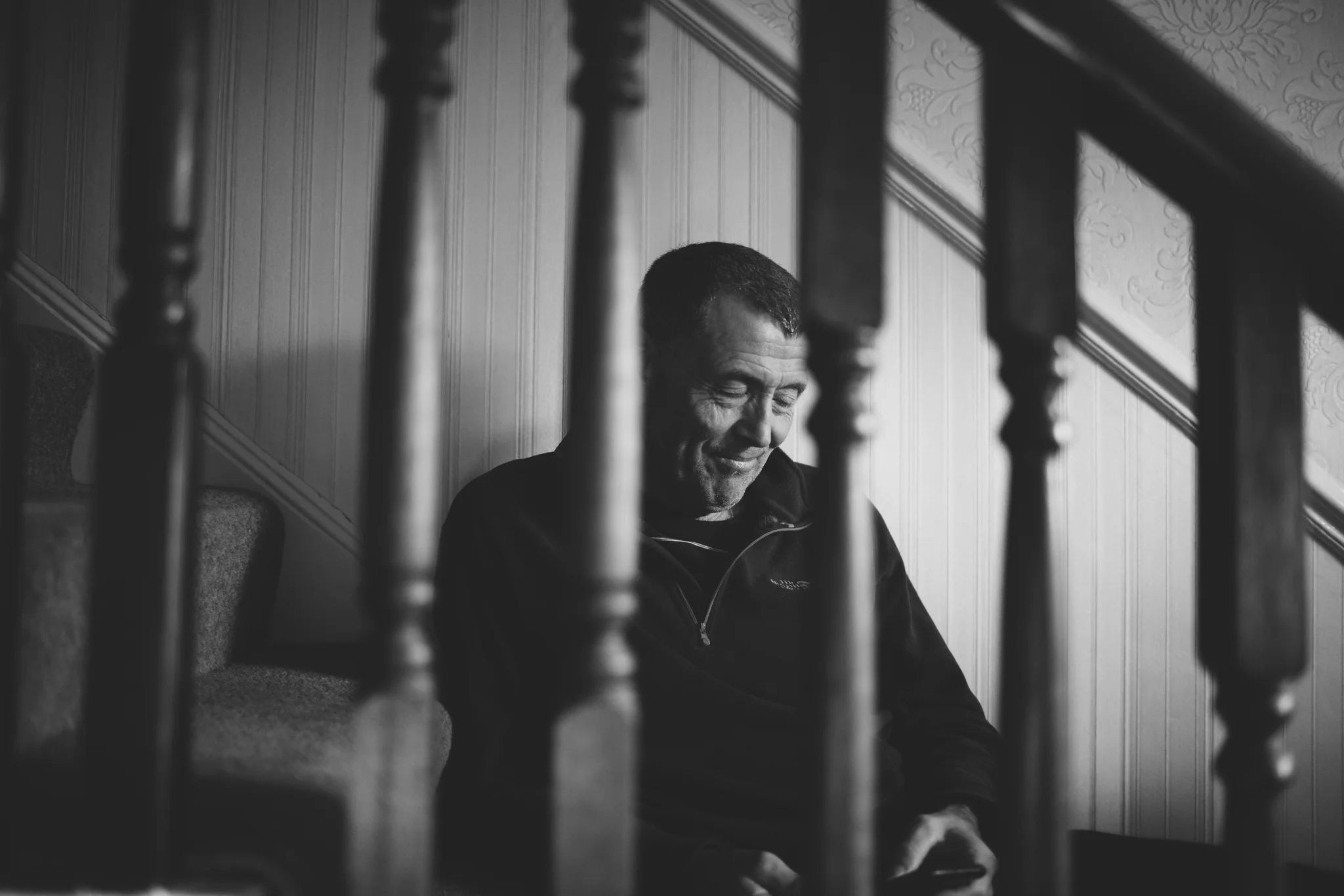 A black and white photo of a man sitting on a staircase, smiling while looking at his phone, framed by the staircase balusters.