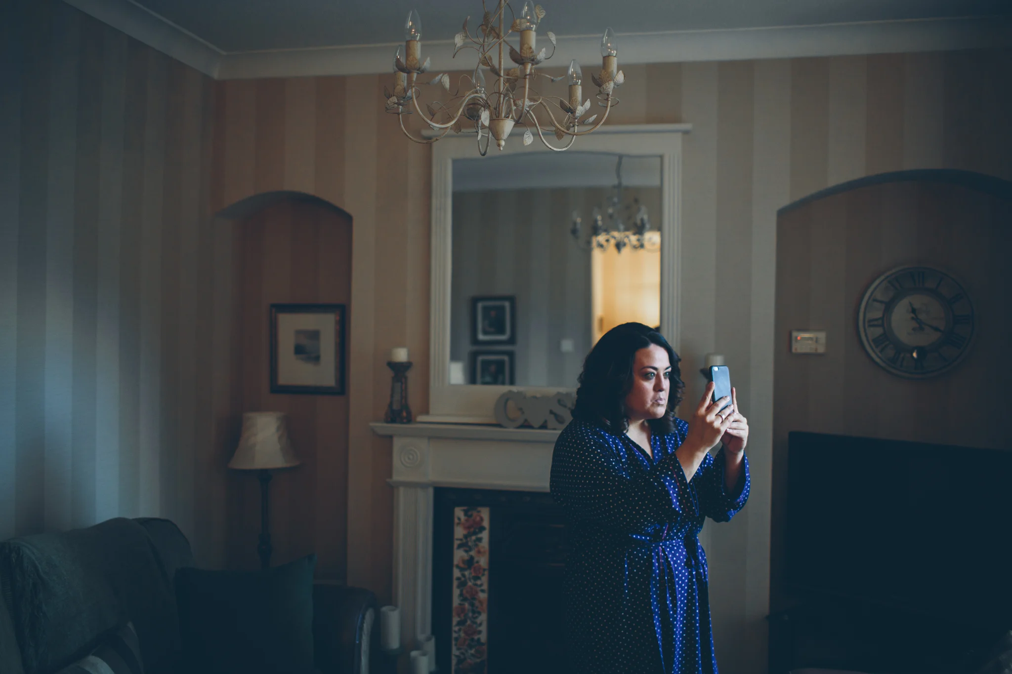 A woman with dark hair wearing a blue polka dot robe stands in a living room, holding a smartphone in front of her face, taking a photo or video.