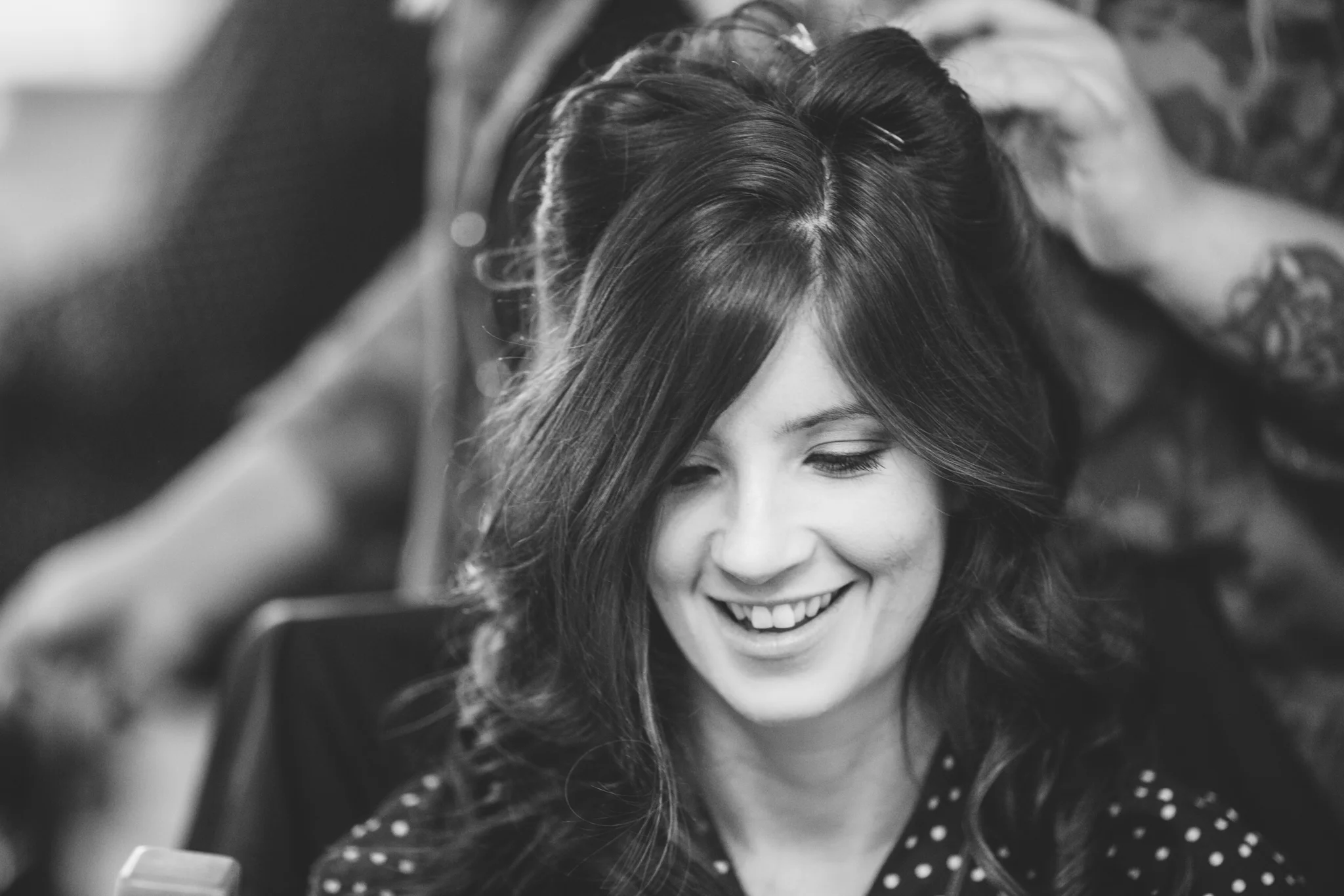 A woman smiling in a black and white photograph, with someone styling her hair.