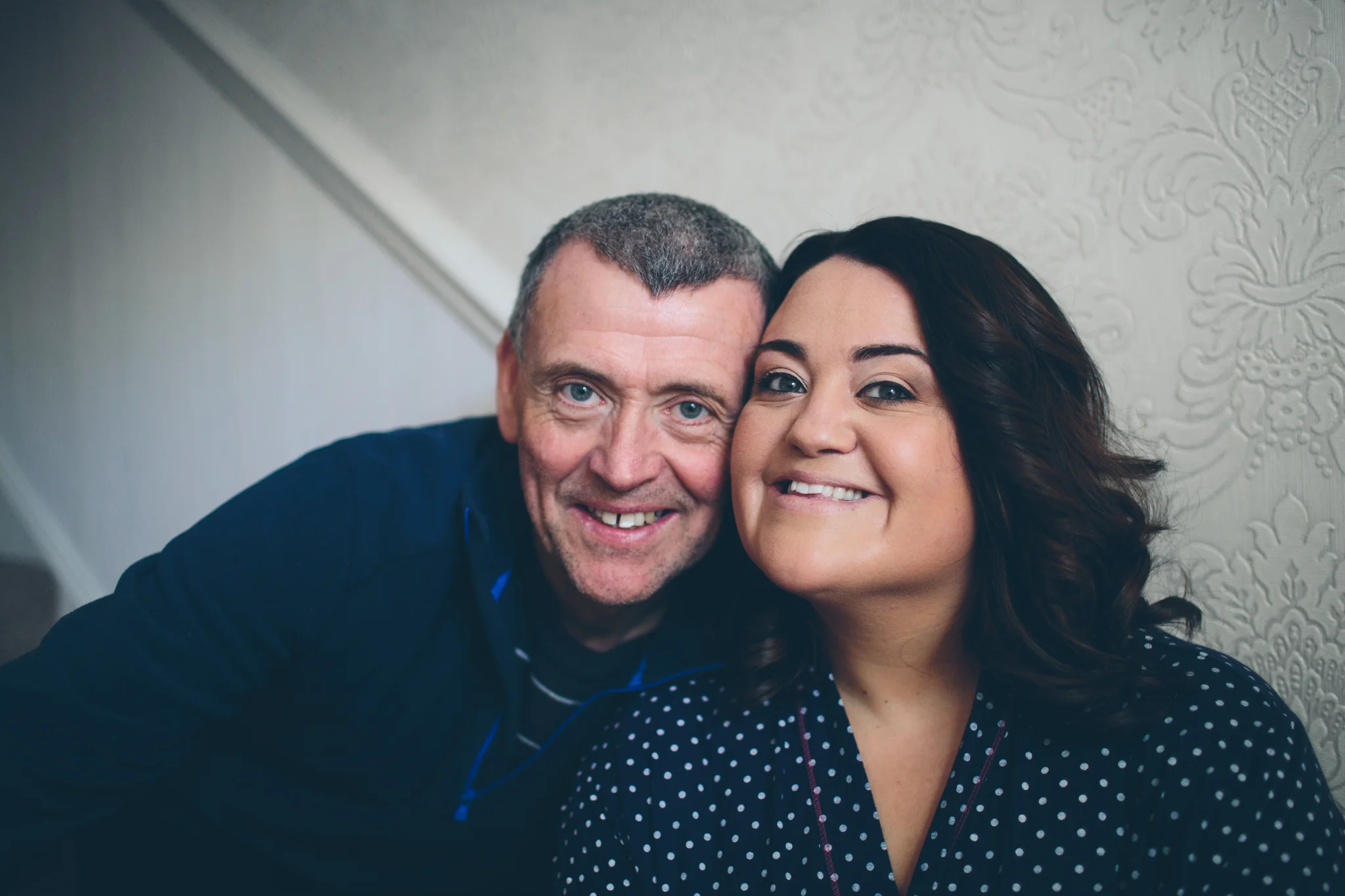 A close-up of a smiling middle-aged man with short hair and a woman with dark brown hair, both showing their teeth, posing together indoors against a textured cream-colored wall.