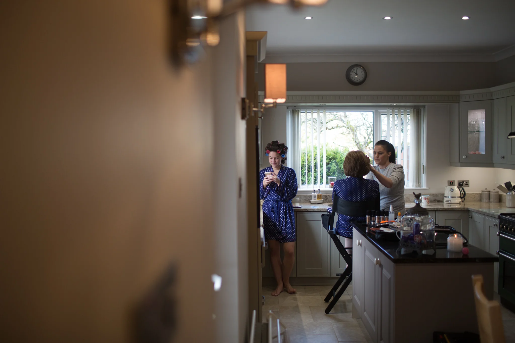 A woman with curlers in her hair sits at a vanity while a makeup artist applies makeup to her face. In the foreground, a woman in a blue polka dot robe is looking at her phone. The kitchen has gray cabinets, a large window with vertical blinds, and v