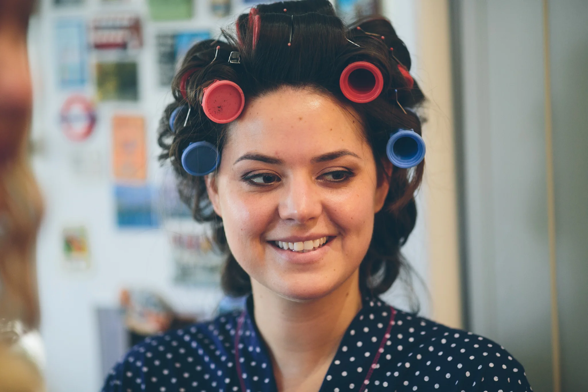 A woman with dark hair in curlers, some pink and some blue, smiling and looking at someone off-camera. She is wearing a dark blue polka dot robe and is in a room with posters and pictures on the wall.