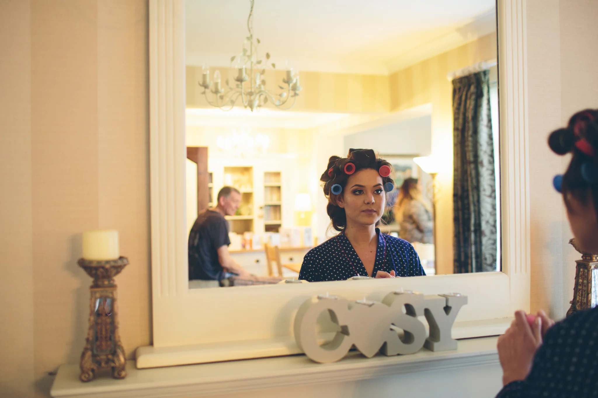A woman with hair rollers in her hair looking at herself in a large wall mirror while getting ready, with the word 'easy' on the shelf below the mirror, in a warmly lit room.